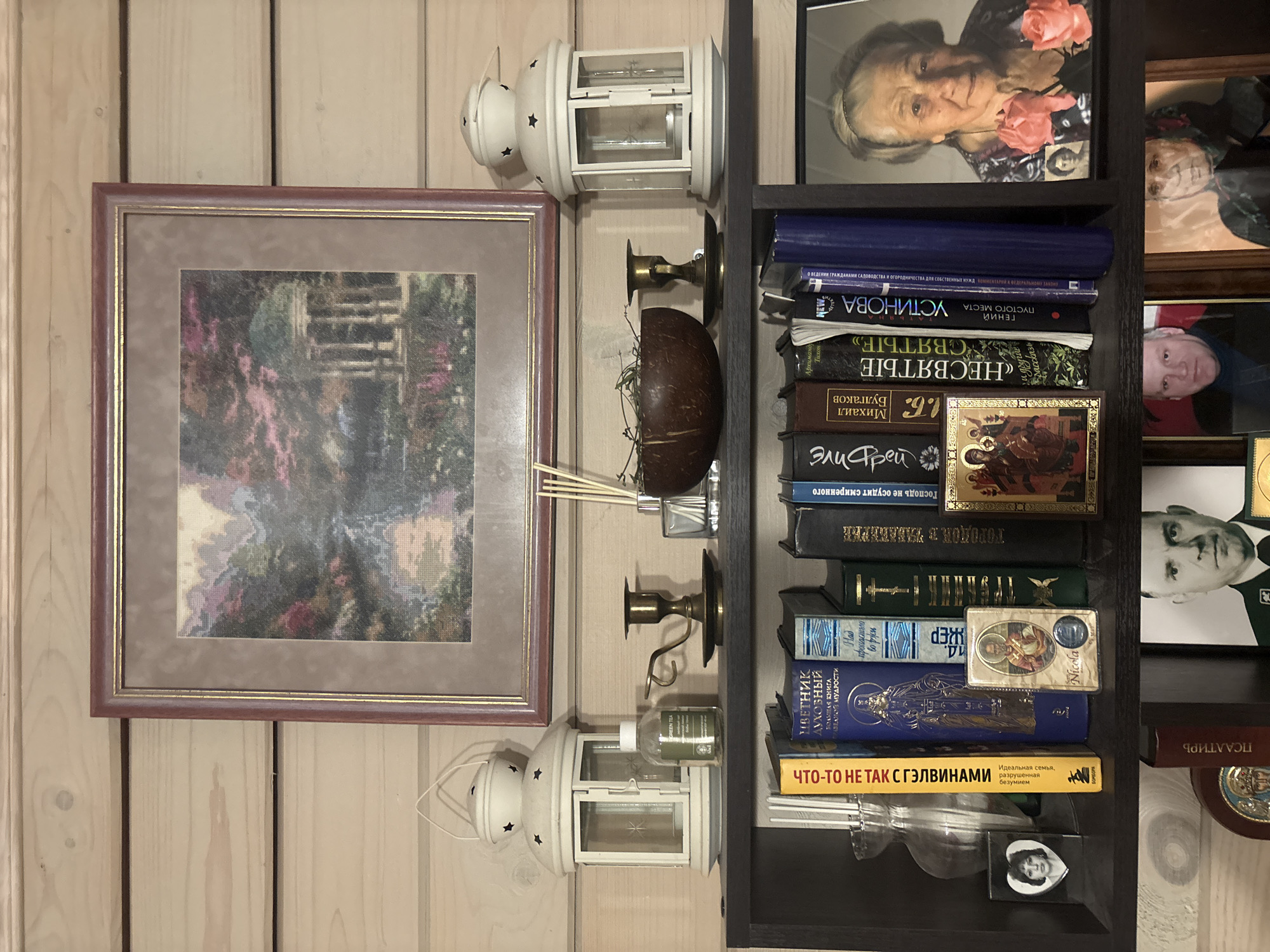 The image depicts a wooden shelf unit with various decorative items and books. At the top, a framed embroidered picture of a landscape with a gazebo is displayed against a wood-paneled wall. Below this, on the shelf, are two white lanterns, a small brass candlestick, a coconut shell bowl with dried twigs, and a bottle of green liquid labeled "GREEN TEA".
The main shelf below holds several books with titles in Cyrillic script, a small heart-shaped locket with a picture of a woman, a small religious icon, and another religious icon with a blue coin. To the right, a framed photograph of an elderly woman with pink roses is placed.
The bottom shelf is partially visible and shows more framed photographs, including one of a man in military or police uniform and another of a woman wearing a headscarf. A book spine with Cyrillic lettering is also visible. The environment appears to be an indoor setting, likely a home, with a rustic wooden aesthetic. The location is identified as Berezkа Snt (Povàrovo), Russia.
