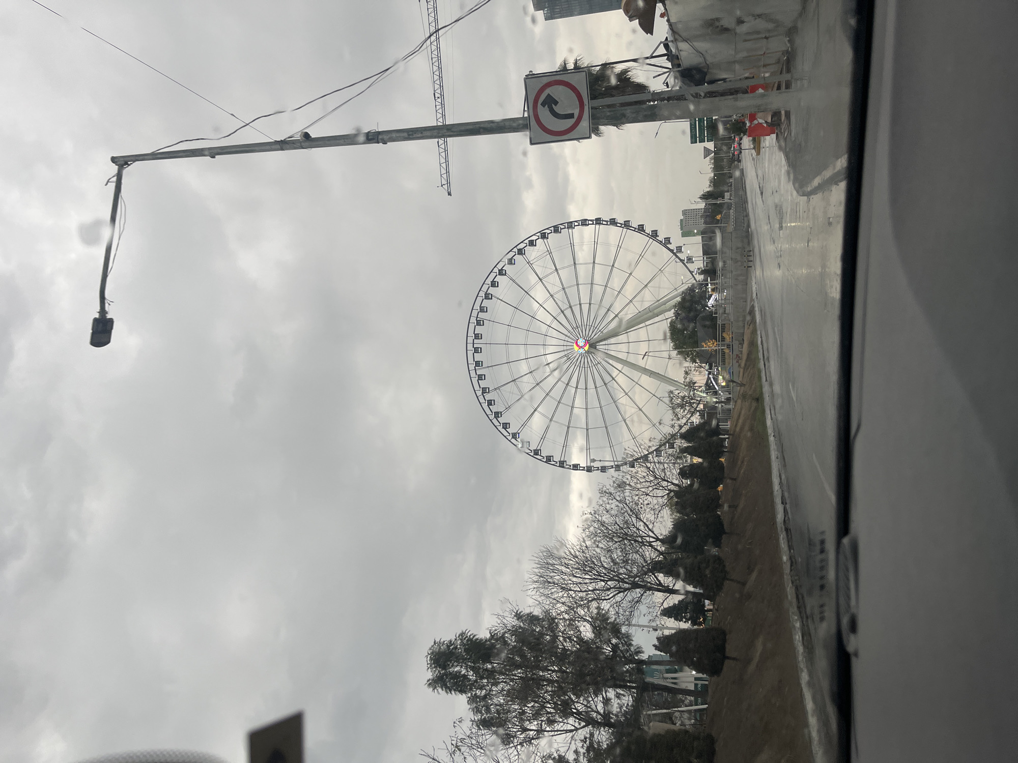 The image shows a view from inside a vehicle, looking out at a street scene in Puebla, Mexico. In the foreground, the edge of the car door or window frame is visible. Beyond that, the street is wet, likely due to recent rain. A large Ferris wheel dominates the middle ground, its white structure contrasting against the overcast sky. Bare trees stand between the Ferris wheel and the road, indicating it may be winter. Streetlights and traffic signs are visible, along with some buildings in the background. A "No U-Turn" sign is attached to a pole. Overall, the scene appears to be a city street with a prominent Ferris wheel feature in Puebla, Mexico.