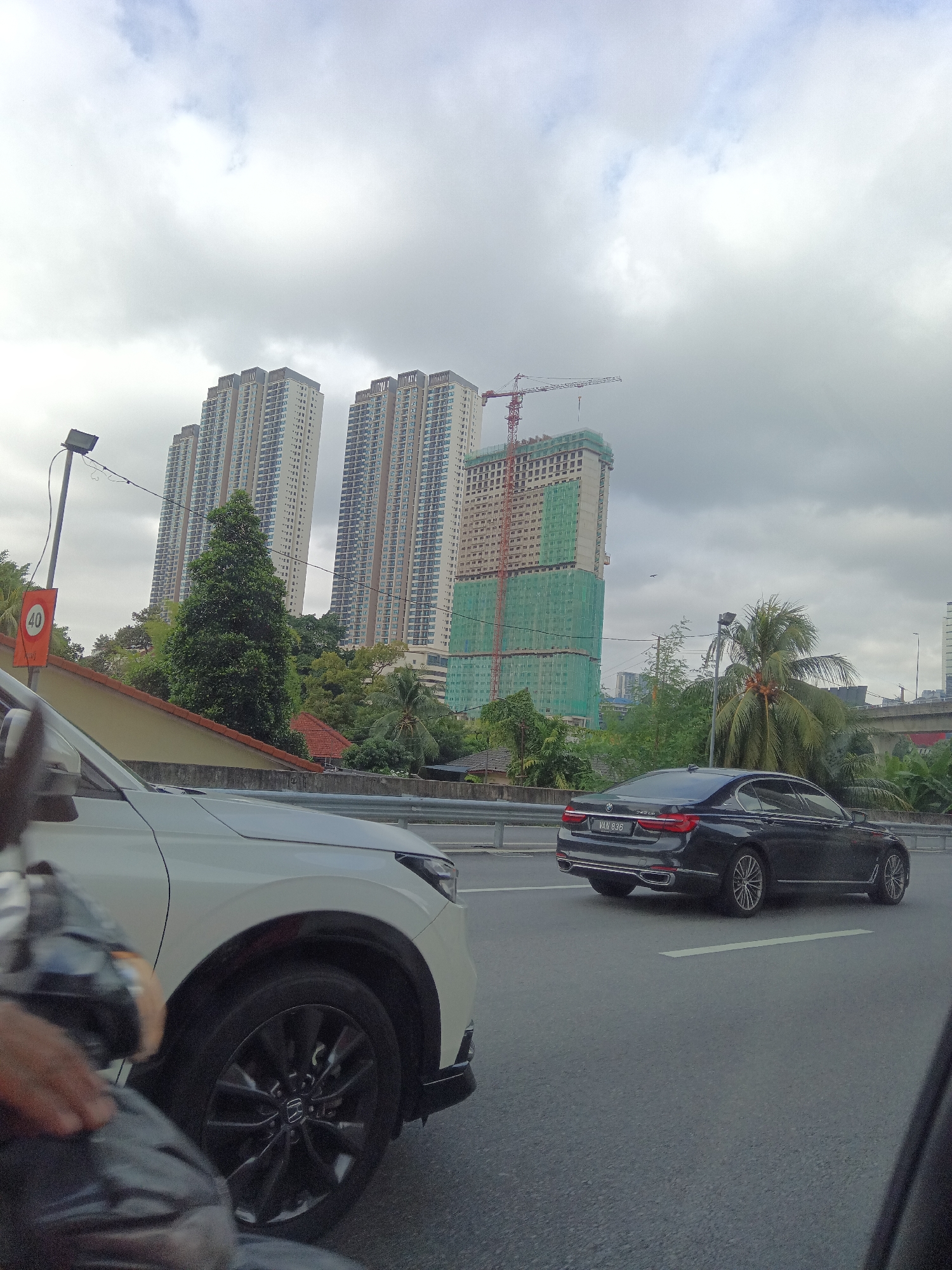 In Kuala Lumpur, Malaysia, on a cloudy day, a black BMW sedan, license plate VAN 836, drives on a multi-lane road. To the left, the front of a white SUV is partially visible. In the background, three tall, modern residential buildings are under construction. One building is covered in green netting, and a red construction crane is visible above it. Lush green trees and foliage surround the buildings. A speed limit sign indicating "40" is mounted on a pole. The road has white lane markings. The scene captures a segment of urban development in Kuala Lumpur.