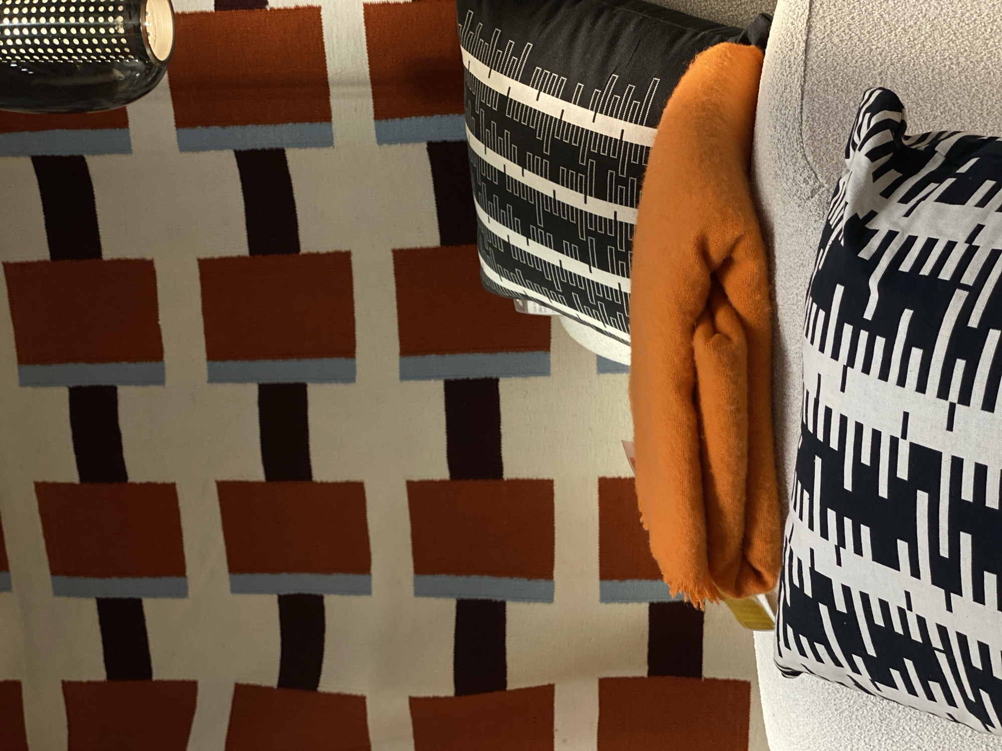 A high-angle, indoor shot focuses on a section of a living room scene. Prominently featured is a rug with a distinctive pattern: rows of rust-colored rectangles are connected by dark brown vertical lines, each rectangle topped by a thin blue-grey horizontal stripe. Positioned on a couch are two decorative pillows: one is black with white geometric lines, and the other is white with black geometric lines. An orange folded throw blanket lies between them. Part of a light fixture with visible holes is visible on the upper left corner. The couch's fabric appears to be light grey. It's impossible to determine the location is San Bernardino Tlaxcalancingo, Mexico or any other geographical location based on the given information.