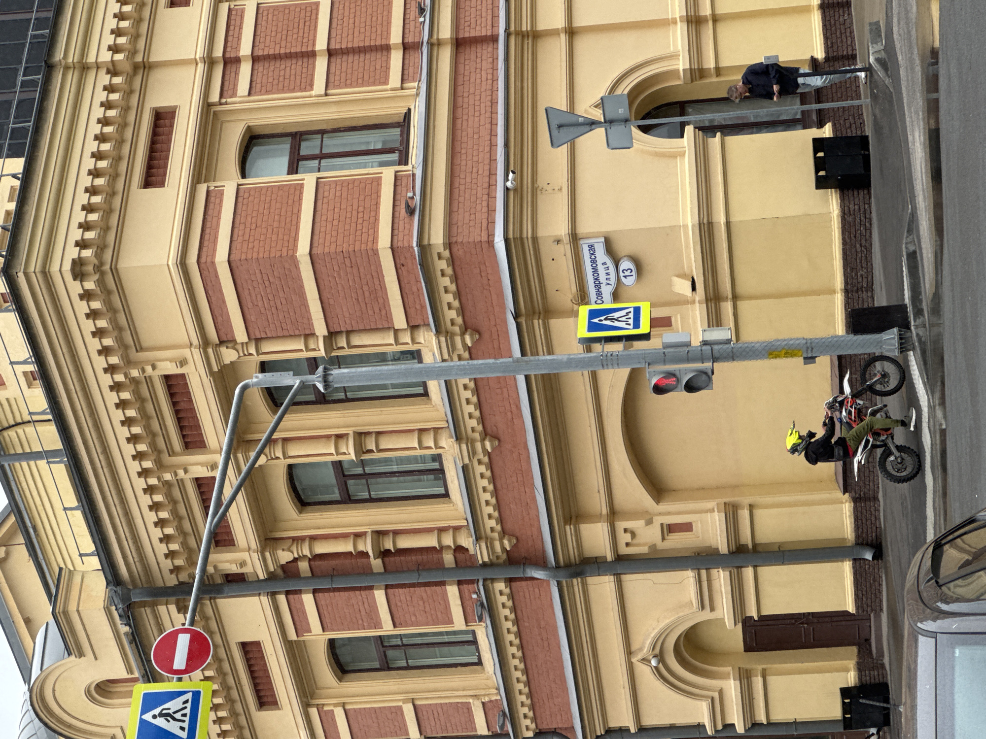 The media file depicts an outdoor scene of a street in Nizhny Novgorod, Russia, with a prominent building and various objects. The building is tall and appears to be an older structure with a decorative facade featuring yellow and red brickwork, arched windows, and elaborate cornices. Several traffic signs are visible, including a pedestrian crossing sign and a circular red and white sign. A traffic light is also present, showing a red signal.
In the foreground, a person is seen riding a dirt bike, wearing a helmet. The bike is angled and appears to be in motion. A car is partially visible on the right side of the frame. Another person is leaning out of an upper window of the building, looking downwards.
The time of day appears to be daytime, and the weather seems overcast. The scene is captured from a slightly low angle, looking upwards towards the building and street elements.
There is visible text on a street sign that reads "Совнаркомовская улица" and a numbered sign "13".