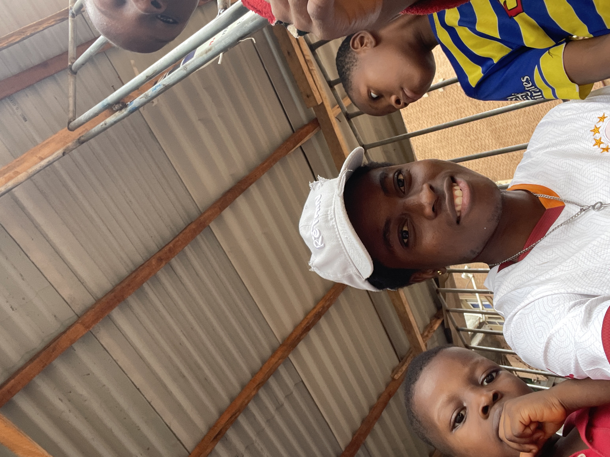 The image depicts four individuals in what appears to be an outdoor setting in Onitsha, Nigeria, likely under a corrugated metal roof structure. A young boy is wearing a yellow and blue striped shirt, and the adult is wearing a white patterned shirt and a light-colored baseball cap. A second young boy is wearing a red shirt. The fourth person is positioned above, partly out of frame. The overall scene seems to capture a casual moment with family.