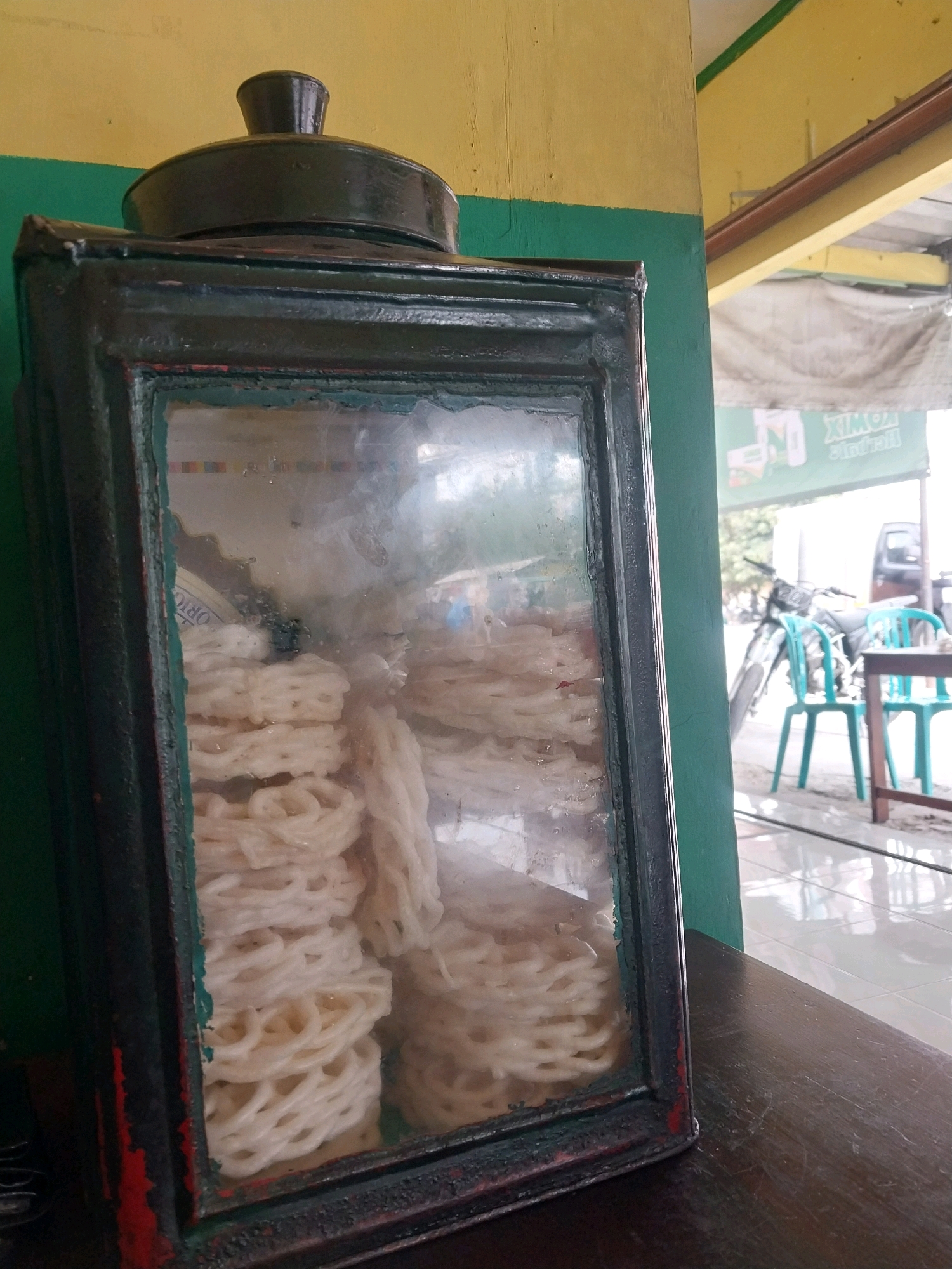 The image shows a close-up of a vintage glass-fronted display case filled with stacks of white, round crackers, likely kerupuk. The case is dark, possibly black or dark green, with visible signs of wear and tear, including chipped red paint on its bottom edge and a slightly rusted metal frame. The display case has a dark, conical lid on top. It sits on a dark, wooden surface, possibly a table, which shows scratches and wear.
Behind the display case, the background is a wall painted in two horizontal bands: a yellow band at the top and a green band below it. To the right of the display case, an open area is visible, suggesting an outdoor or semi-outdoor setting, perhaps a warung or food stall. In this background area, there are several bright teal plastic chairs arranged around a wooden table. A motorcycle is parked nearby. A banner with Indonesian text, including the word "XIMON," is visible hanging in the background. The flooring appears to be tiled. The scene is likely located in Sayegan, Indonesia, as indicated by the presence of typical Indonesian food items and the architectural style. The lighting suggests it is daytime.