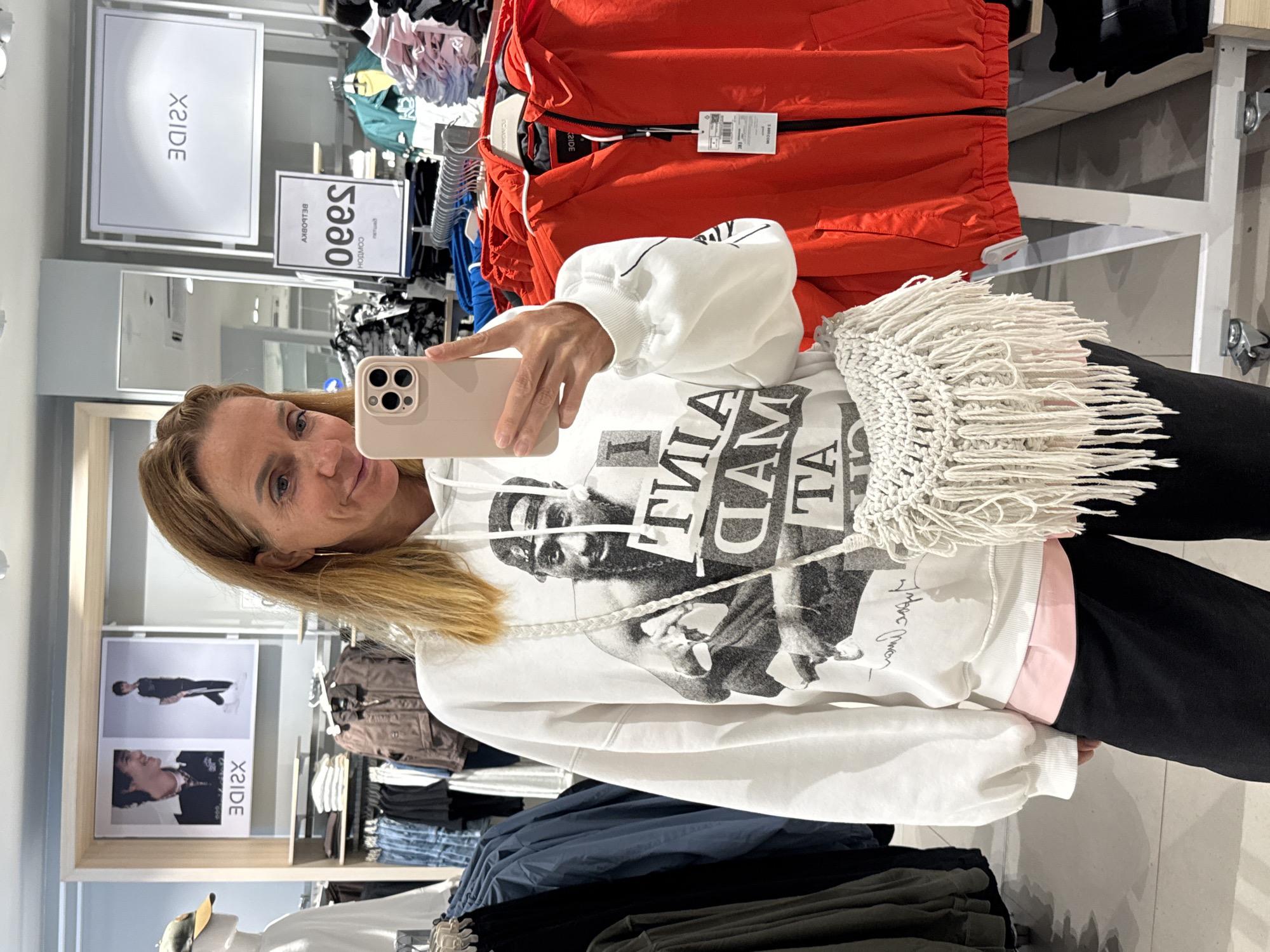 A woman takes a selfie in a clothing store in Bishkek City, Kyrgyzstan, with the image rotated 90 degrees counter-clockwise. The woman, with shoulder-length blonde hair, appears in the foreground, looking at the camera with a slight smile. She holds a light beige smartphone, visible in the center-left, with which she is taking the photo.
She is wearing a white oversized hoodie with a prominent black and white graphic print on the front, depicting a person's face and upper body, overlaid with large, bold, fragmented text, part of which reads "I AM A MAD T". A light pink layer is visible underneath the white hoodie at her waist, and she is wearing dark-colored pants. A distinctive, cream-colored, fringed macrame-style bag hangs from her shoulder, positioned on her right side.
The background clearly indicates an indoor retail setting. Racks of clothing are visible throughout, with bright red jackets on hangers occupying the right side of the frame. Other garments in various colors, including blues, blacks, and whites, are displayed on shelves and hangers. Several white rectangular signs are present; one large sign prominently displays "X2IDE" (reversed text), and a smaller one below it shows reversed Cyrillic-like text, including "БЕЛВОРУ", likely a brand or product name. Another sign on the far left also features "X2IDE" (reversed) alongside images of models. The top of a mannequin wearing a cap is partially visible in the lower left corner. The scene captures a casual moment in a bustling store environment.