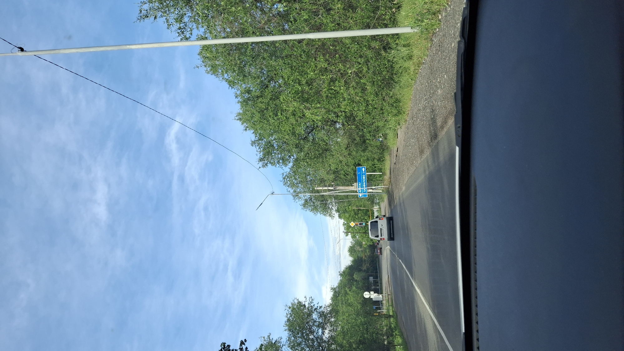 View from a vehicle driving on a road in or near the city of Түзусай, Kazakhstan. The sky is blue with clouds. Utility poles run along the side of the road. Trees line the side of the road. A road sign indicates the direction to Матай and Панфилово. A white van drives on the road. A traffic light is visible.