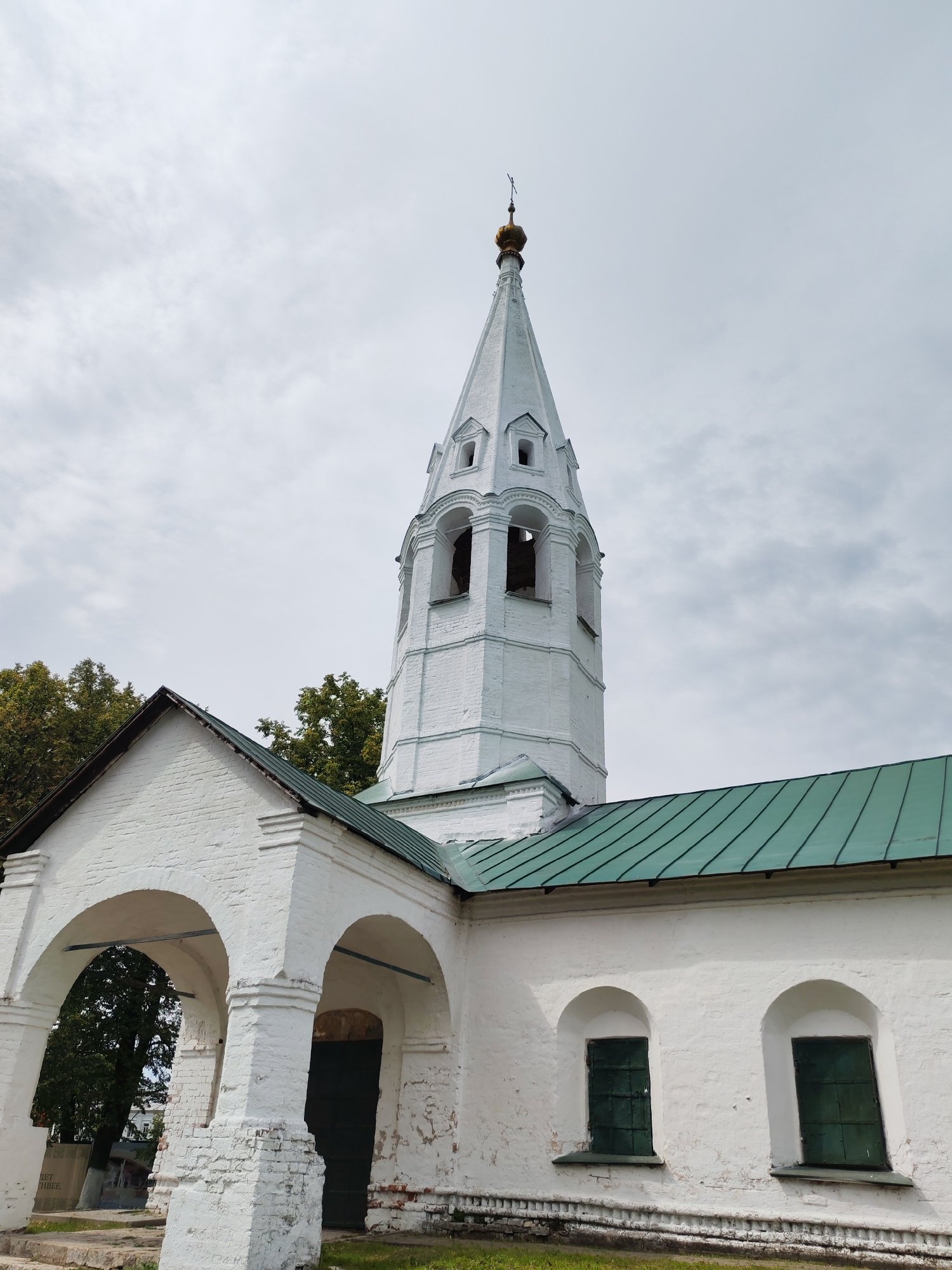 The image captures a white brick church in Rusanovo, Russia, under a cloudy sky. The main structure features a tall, octagonal bell tower topped with a golden dome and a cross. The tower has arched openings for bells and decorative architectural details. Attached to the bell tower is a lower building with a green metal roof, a white brick facade, and arched entrances. Two arched windows with dark green shutters are visible on the side of this lower building. In the background, mature trees with green foliage can be seen. The ground in front of the building is covered with grass, and there is a stone pathway leading to the arched entrances.