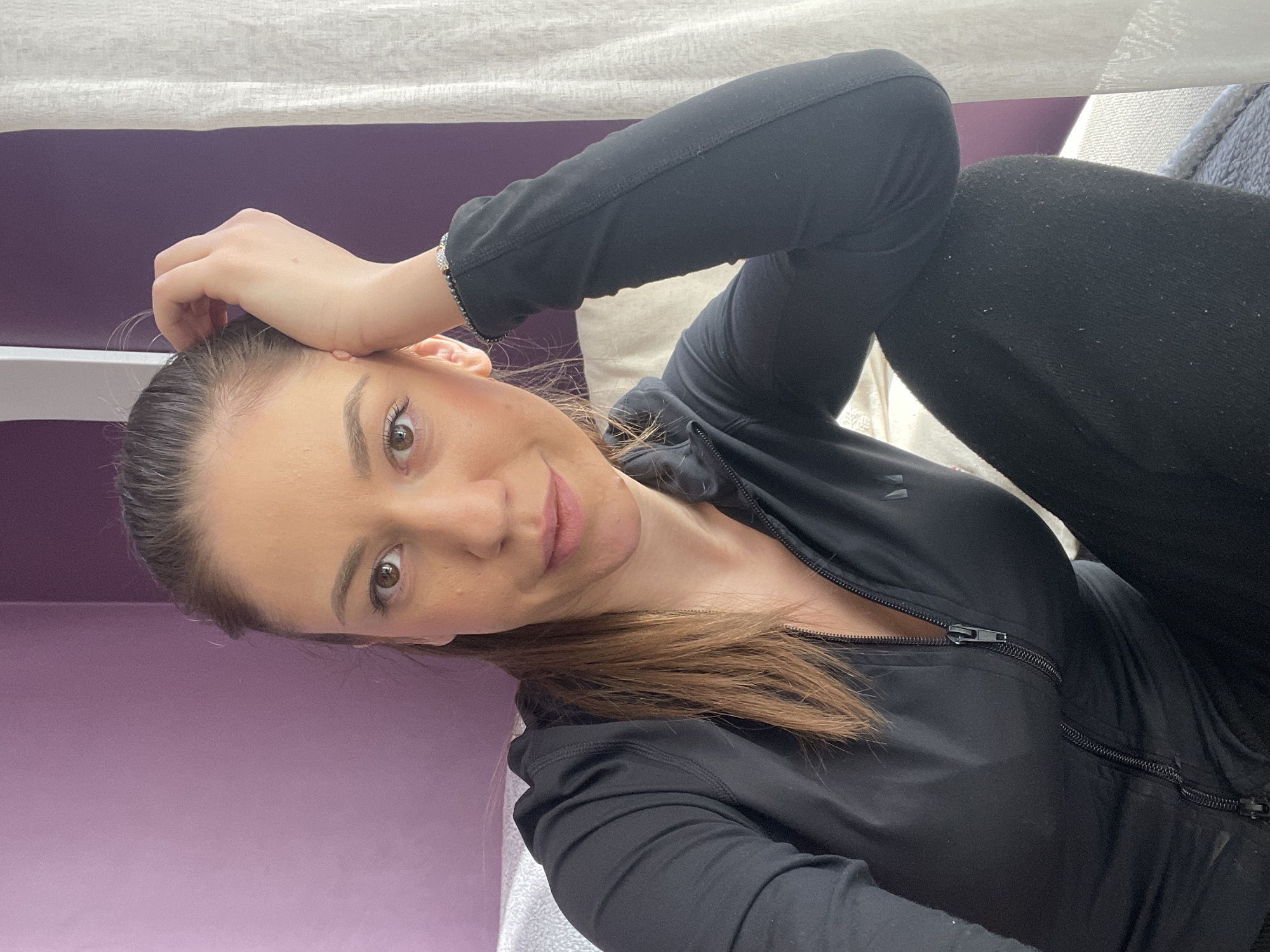 A young woman is looking directly at the camera while lying on her back. She has medium-length brown hair. She is wearing a black zip-up jacket and black leggings. She is lying on a white pillow or cushion, and behind her, there is a purple wall. She has one hand on her head. The location is not discernible from the image; therefore, it is impossible to ascertain that the location is Sofia, Bulgaria.
