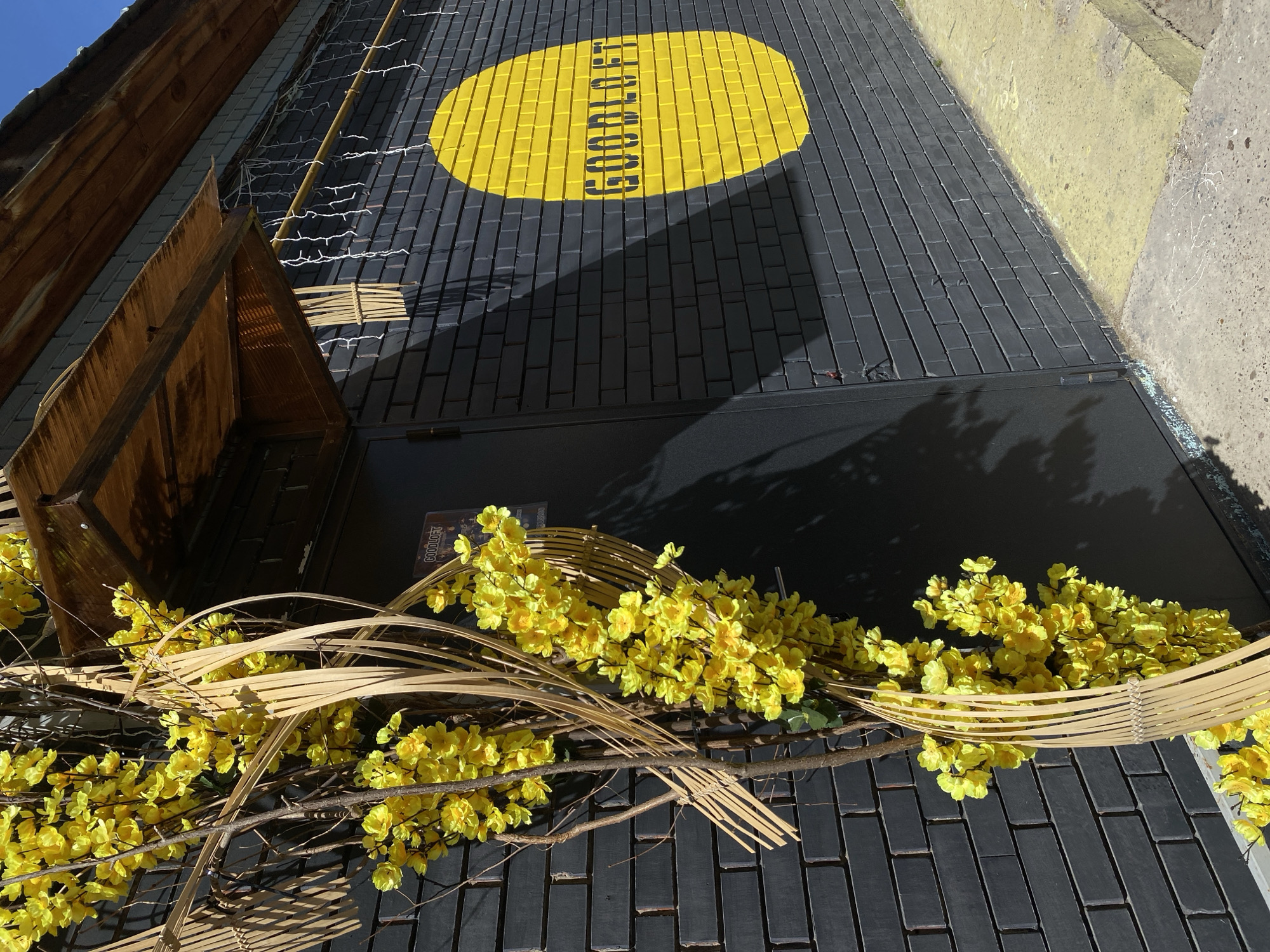 The image captures the exterior of a building in Votkinsk, Russia. The facade is constructed from dark bricks, arranged in a linear pattern. A prominent yellow oval shape is painted on the brick wall near the top of the building, with "GOOD LOFT" stenciled in black within the oval. The text is oriented vertically. The bottom half of the image is dominated by artificial yellow flowers and woven bamboo elements that seem to be decorative. The building's entrance appears to be a simple, dark door. The image exhibits shadows that indicate sunlight is present. In the upper left corner, wooden architectural features are visible, perhaps a part of the building's design or a sign structure. The overall setting seems to be an urban area.