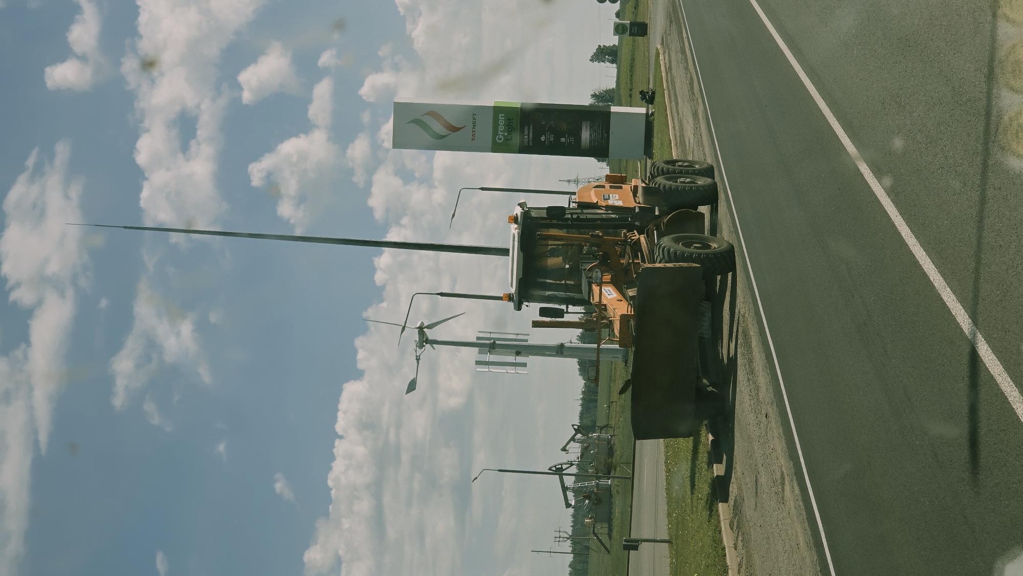 An orange grader is parked on the shoulder of a paved road. The grader has a large blade at the front and large tires. Behind the grader, to the left, are several oil derricks and a small wind turbine. To the right of the grader is a tall, slender pole topped with a pointed object. Further to the right is a large vertical sign for "TATNEFT Green Light" which appears to be a gas station. The sky above is blue with scattered white clouds. The scene appears to be in a rural or industrial area.