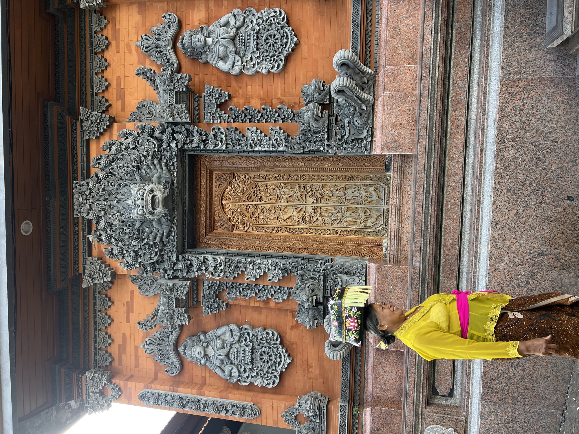 A woman in traditional Balinese clothing stands in front of a highly decorated Balinese temple doorway in Singakerta, Indonesia. The doorway features intricate carvings in brick and stone, including figures and floral patterns. The woman wears a bright yellow lace top, a brown batik skirt, and a floral headpiece. A pink sash is tied around her waist. She appears to be smiling. The doorway itself is made of carved wood. The wall surrounding the doorway is reddish-brown and features gray stone carvings. The scene is brightly lit.