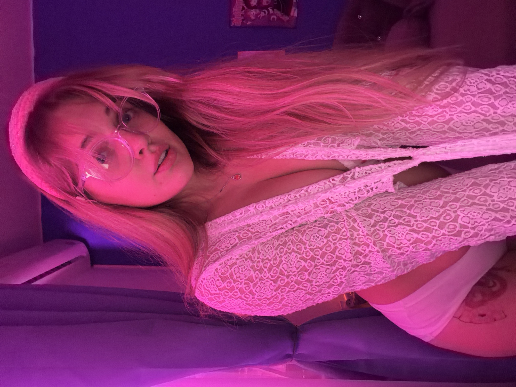 A young woman with long pink hair and eyeglasses is depicted in a selfie-style image. She is wearing a white lace top and white underwear. A floral tattoo is visible on her thigh. The environment appears to be indoors, with a blue wall and a headboard in the background. A photograph is visible on the wall. The image is heavily illuminated with pink and purple lighting. The location cannot be independently confirmed.