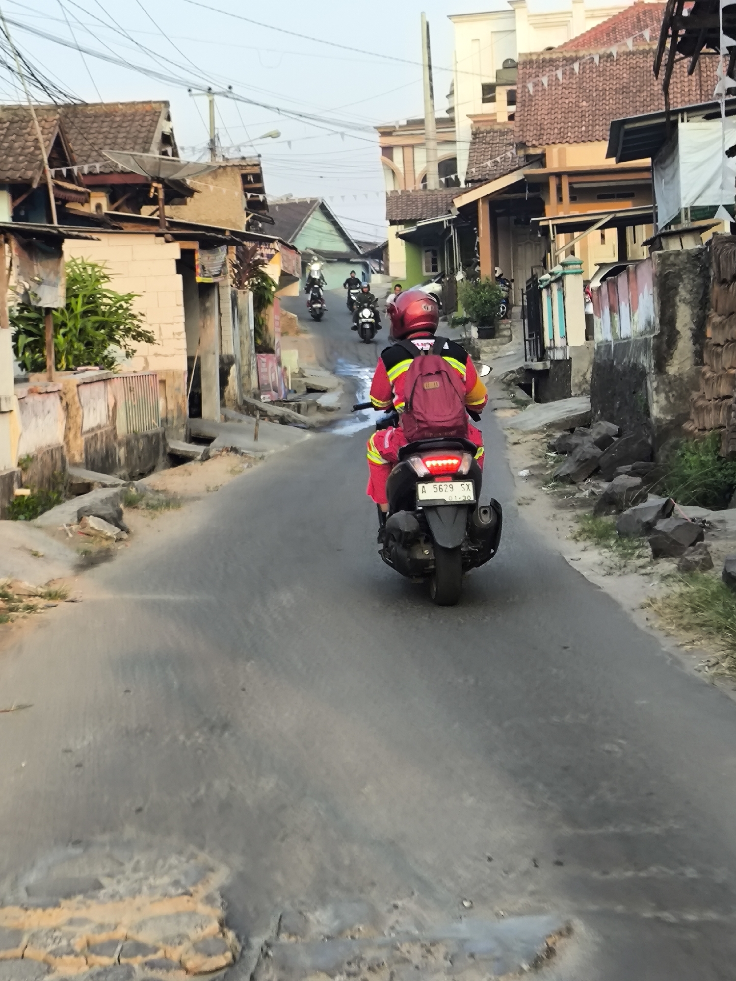 In Cilegon, Indonesia, a motorcycle rider wearing a red helmet and a red and yellow jacket is seen from behind, riding on a narrow, uneven asphalt road. The rider has a red backpack on their back. The road is flanked by houses with tiled roofs and some vegetation. Other motorcycles are visible in the distance further up the road. The environment appears to be a residential area with overhead electrical wires creating a complex network.