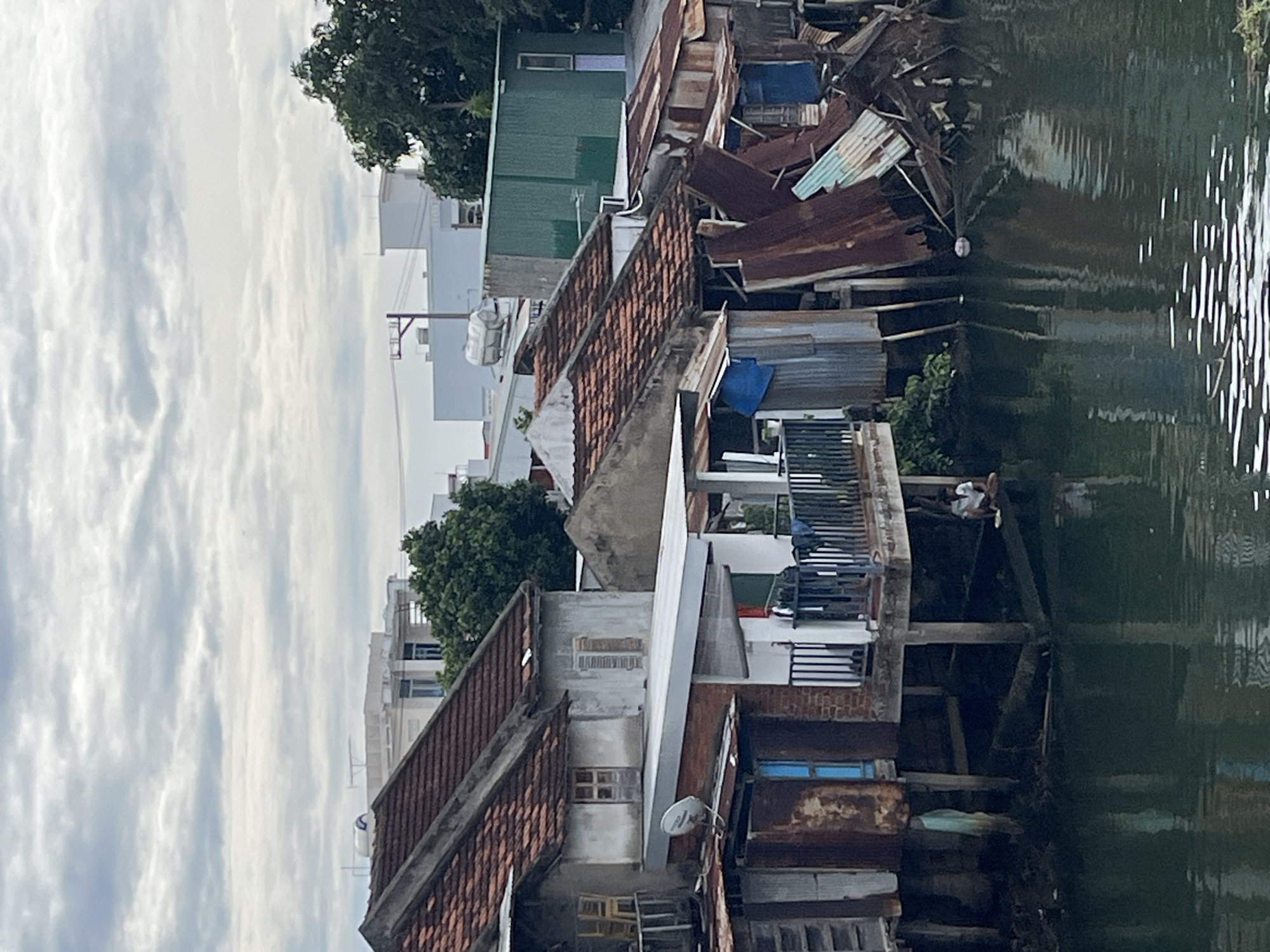 This image captures a waterside scene in Nha Trang, Vietnam, showing a row of weathered, stilted structures lining a canal. The buildings, constructed from a mix of corrugated metal, wood, and concrete, appear to be in a state of disrepair, with rusted metal siding and some structural damage. The roofs are predominantly tiled, some with evidence of wear.
A person is seated on a wooden beam extending from one of the structures over the water, appearing to be engaged in some activity not clearly discernible. Clothes are visible hanging on a railing on one of the balconies. The canal itself is dark and reflects the overcast sky and the surrounding buildings. In the background, more modern buildings and trees suggest a contrast between older and newer urban development. The sky is cloudy, indicating a diffused light condition. The overall impression is one of a densely populated, possibly informal, residential area situated along a waterway.