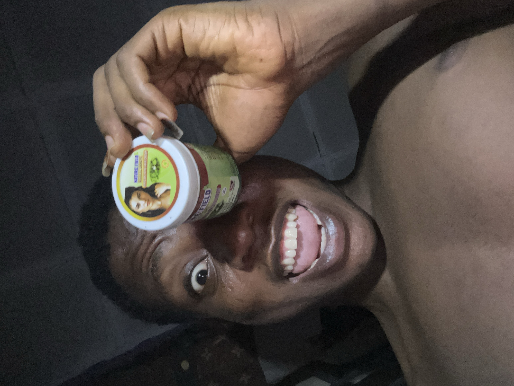 A close-up, angled shot depicts a person holding a small jar of "NATURE'S FIELD" product near their eye. The person is smiling widely, exposing their teeth. The background is dark and indistinct. The location is unknown.