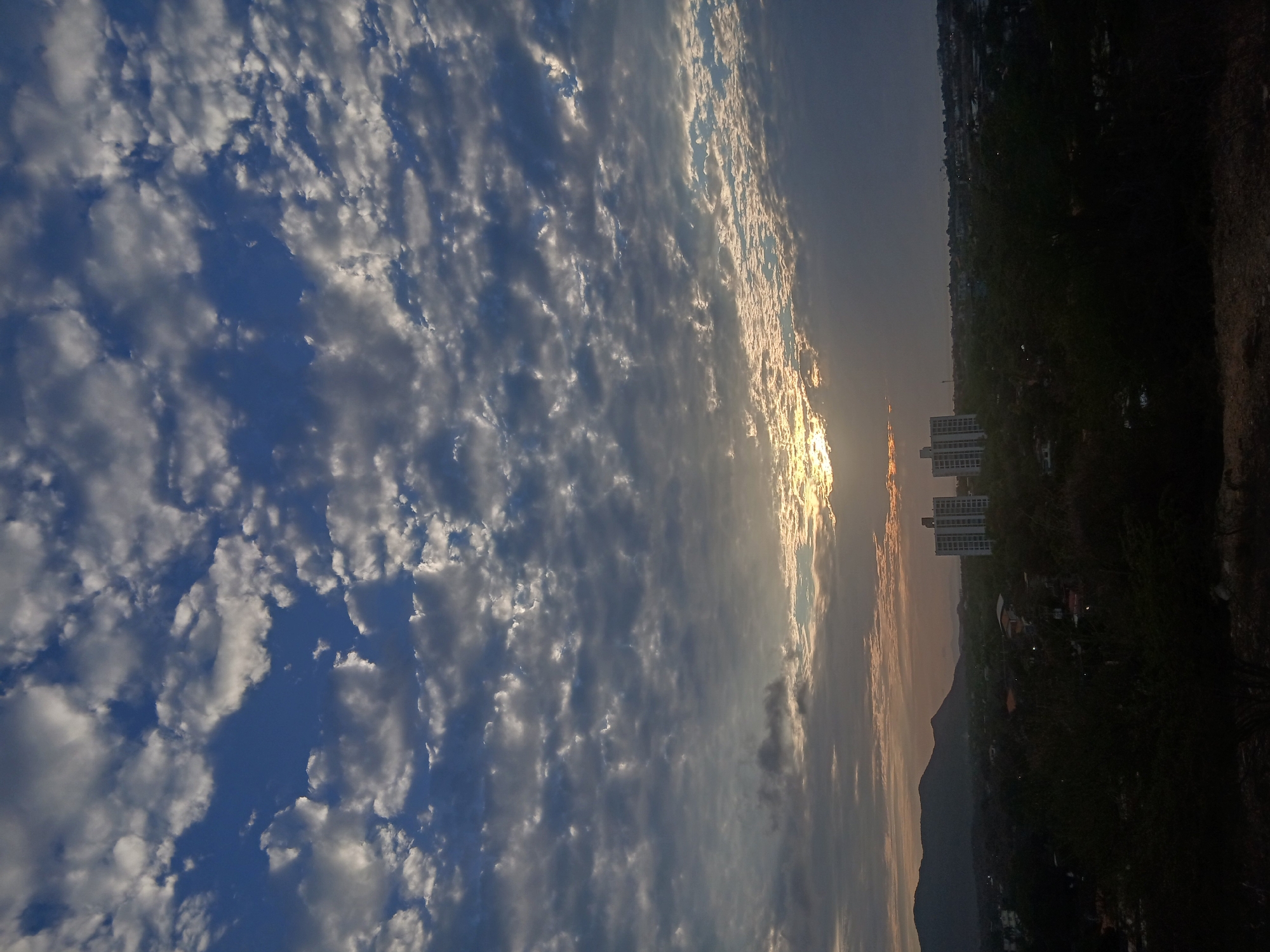 The image shows a cityscape at sunrise or sunset in Juan Griego, Venezuela. The right side of the frame is dominated by dark, silhouetted hills and vegetation, with buildings interspersed. A distant mountain peak is visible. Three apartment buildings stand out, their facades reflecting the sunlight. The left side is filled with a sky containing clouds, creating a contrast in illumination. The sun is located on the horizon, obscured by the clouds.