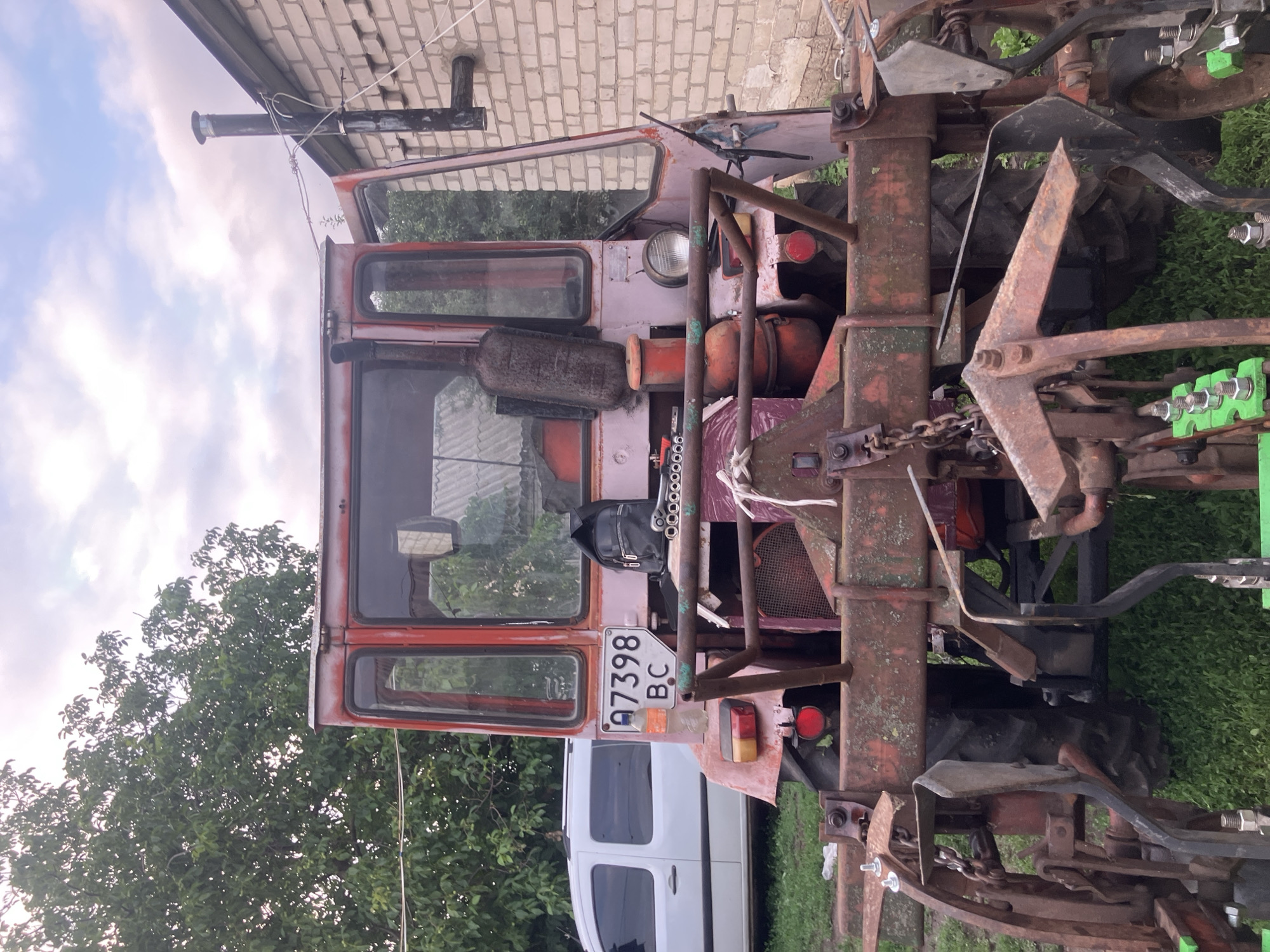 The image depicts the rear view of a weathered, pink and red tractor parked outdoors on a cloudy day in Vovkivka, Ukraine. The tractor, which appears to be agricultural equipment, has a visible license plate reading "A7398 BC". Attached to the rear of the tractor is farming implement, likely a plow or seeder, with various metal components. To the left of the tractor, a white van is partially visible. Behind the tractor, a brick building with a dark exhaust pipe extending upwards is present, and further back, trees and foliage are seen. The ground is covered in grass. The overall scene suggests an outdoor, rural or semi-rural setting, with the tractor appearing to be in use or stored.