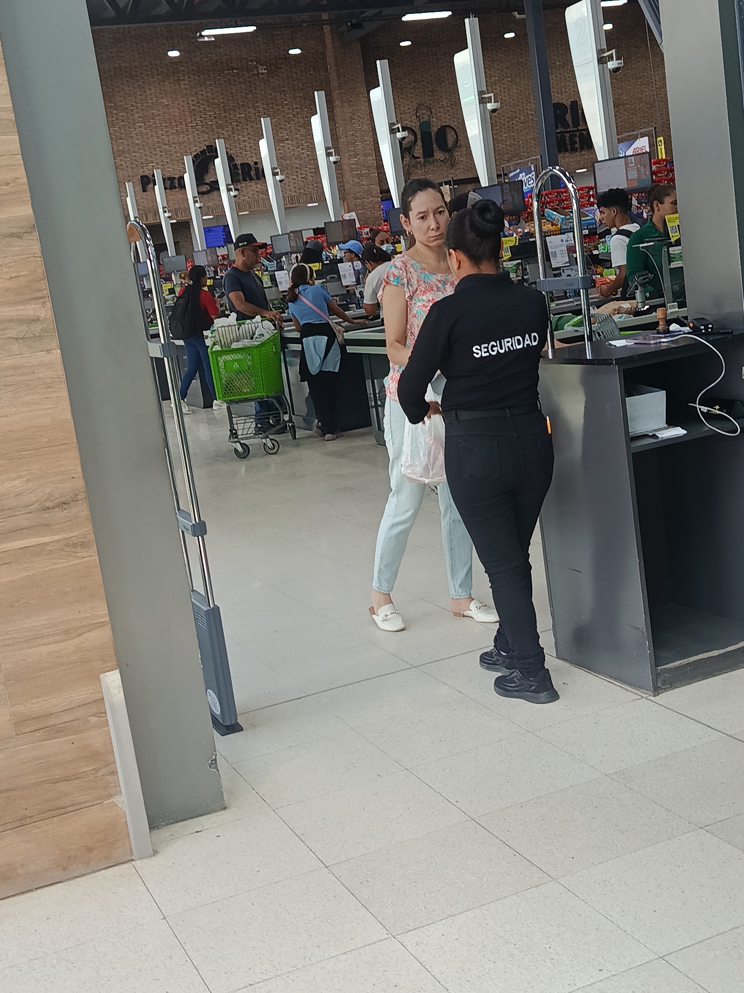 The image captures an indoor scene inside a supermarket, specifically near the checkout and exit area.
In the foreground, two women are prominent. One woman, with dark hair tied back, is seen from the back, wearing a black long-sleeved uniform shirt with the word "SEGURIDAD" (Spanish for "SECURITY") in white letters across the upper back, and black pants and sneakers. She is facing another woman, who is seen from the front. This second woman has dark hair and is wearing a light-colored, short-sleeved top with a floral pattern, light-colored pants, and white slip-on shoes. She holds a translucent white plastic bag in her left hand and has a neutral to slightly serious expression as she looks at the security guard.
Behind these two women, a bustling supermarket environment is visible. Several checkout counters are active, with cashiers attending to customers. One customer, a man in a black cap and dark t-shirt, is pushing a bright green shopping cart. Another customer, wearing a blue shirt, is also at a register. Shelves stocked with various products can be seen behind the registers. The background features a brick-patterned wall, and parts of large signs or decorations with the text "Plaza Rio" are visible.
To the left, a metal security gate or scanner is positioned at the exit point. The flooring transitions from light-colored square tiles in the main store area to a wooden-patterned floor near the exit. The scene appears to be well-lit by artificial lighting, typical of a retail establishment. No specific time of day or outdoor weather can be determined.
