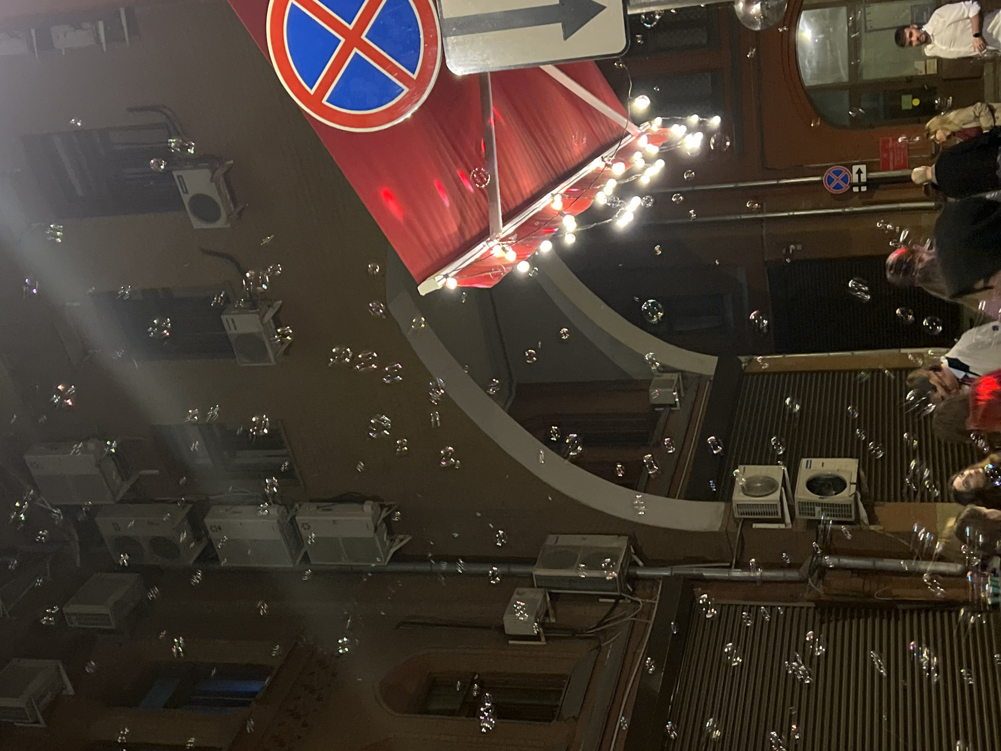 The image captures a city scene at night, filled with soap bubbles. Buildings form the backdrop with multiple air conditioning units attached to the walls. A red awning decorated with lights is visible on top of one of the buildings. A "No Parking" sign with an arrow points to the right. People are present on the right-hand side, although their faces are partially obscured.