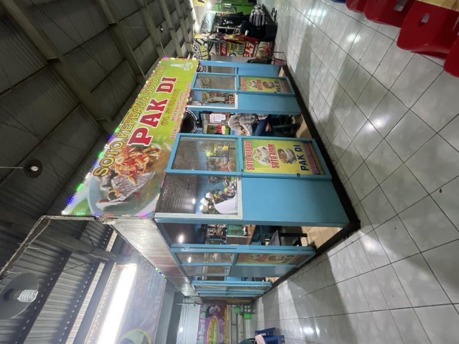 The scene depicts an indoor food court or market stall, specified to be located in Nganguk, Indonesia. The central subject is a rectangular, light blue food stall labeled "SOTO KERBAU & SOTO AYAM PAK DI (H. SUMADI)". The stall features light blue solid panels on the lower half and clear or mesh panels on the upper, with a dark base indicating a raised floor. A large green and yellow banner displaying the stall's name, an image of soto, and text including "BLOK D LANTAI 2 PASAR KLIWON KUDUS" is affixed to the top, illuminated by multi-colored LED strip lights. Inside the stall, a person in a white shirt and cap stands behind a counter on the left. Another individual, wearing a headscarf and light-colored top, is seated at a table inside on the right. The floor consists of light grey square tiles. The overhead structure features a high, corrugated metal roof supported by metal beams, from which various fluorescent tube and incandescent lights are suspended. Stacked red plastic stools are visible in the right foreground. To the far left, a person's lower body is visible seated on a blue stool, adjacent to another partially visible banner. Other commercial stalls are discernible in the right background.