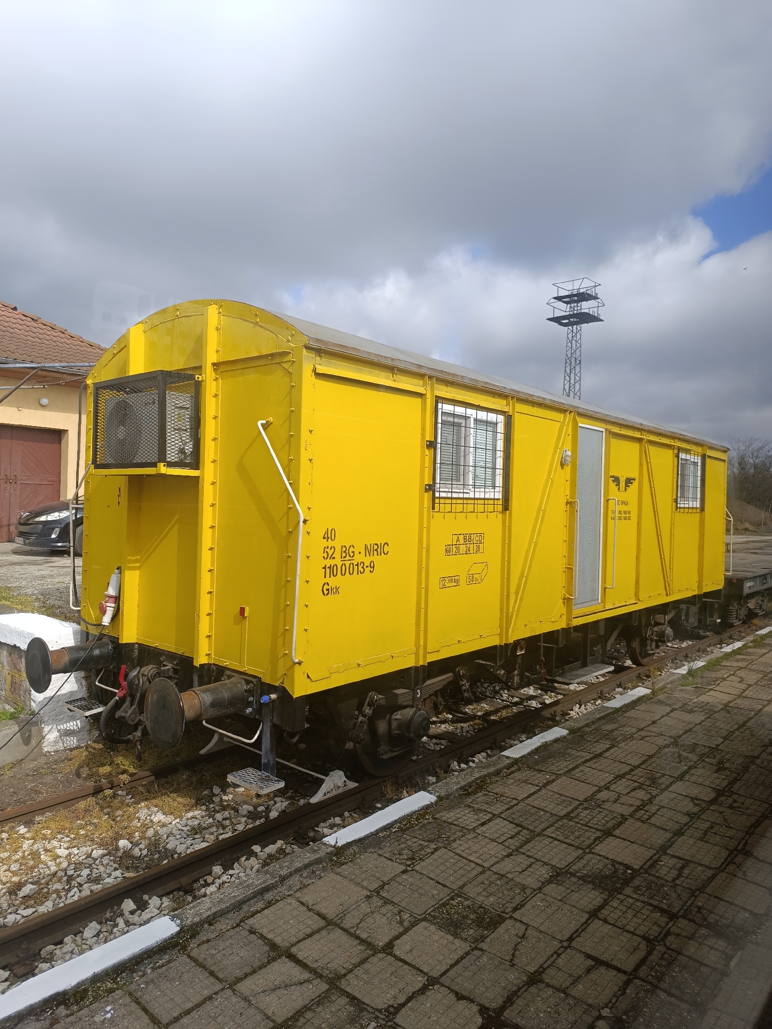 The image shows a yellow railway wagon, possibly in the city of Unknown, Unknown. The wagon is sitting on railroad tracks next to a paved area. It has a number of windows and a door, all with metal bars. On the side of the wagon, there are various markings and numbers. The wagon appears to be in a rail yard or siding. In the background, there's a building, a car, and a metal tower. The sky is overcast.