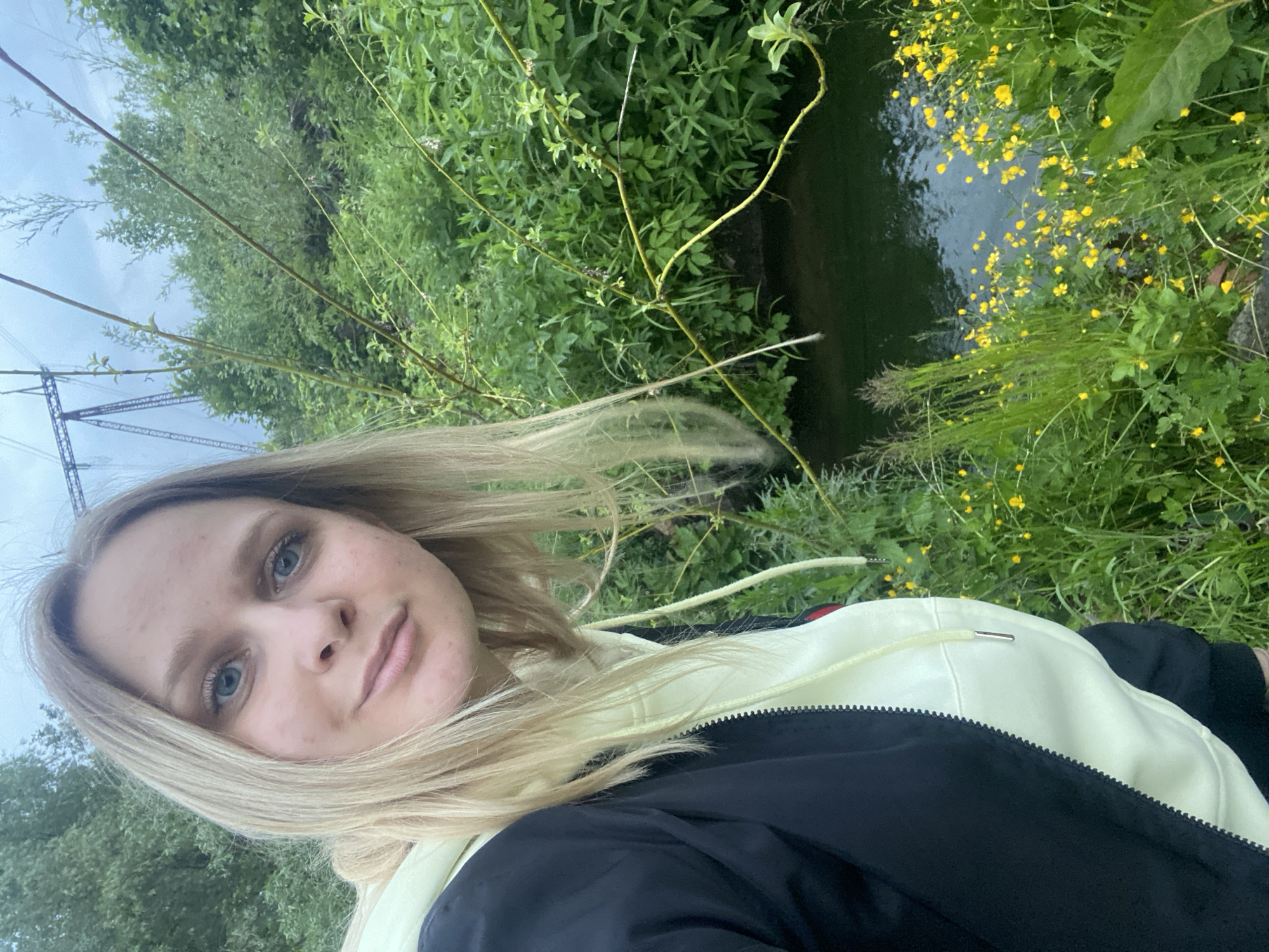 A young blonde woman is centered in a close-up selfie-style shot, looking directly at the camera with a neutral expression, against a natural outdoor backdrop in the city of Unknown, Unknown. She has blue eyes and her hair falls to her shoulders. She wears a black zippered jacket over a light yellow hoodie with a visible drawstring. The background features lush green foliage and dense vegetation, including a small body of dark green water to the lower right. Numerous small yellow flowers are scattered among the green plants on the bank. In the upper right quadrant, a large metal power line transmission tower is partially visible against a light, overcast sky. Thin tree branches extend from the left towards the center and right foreground.