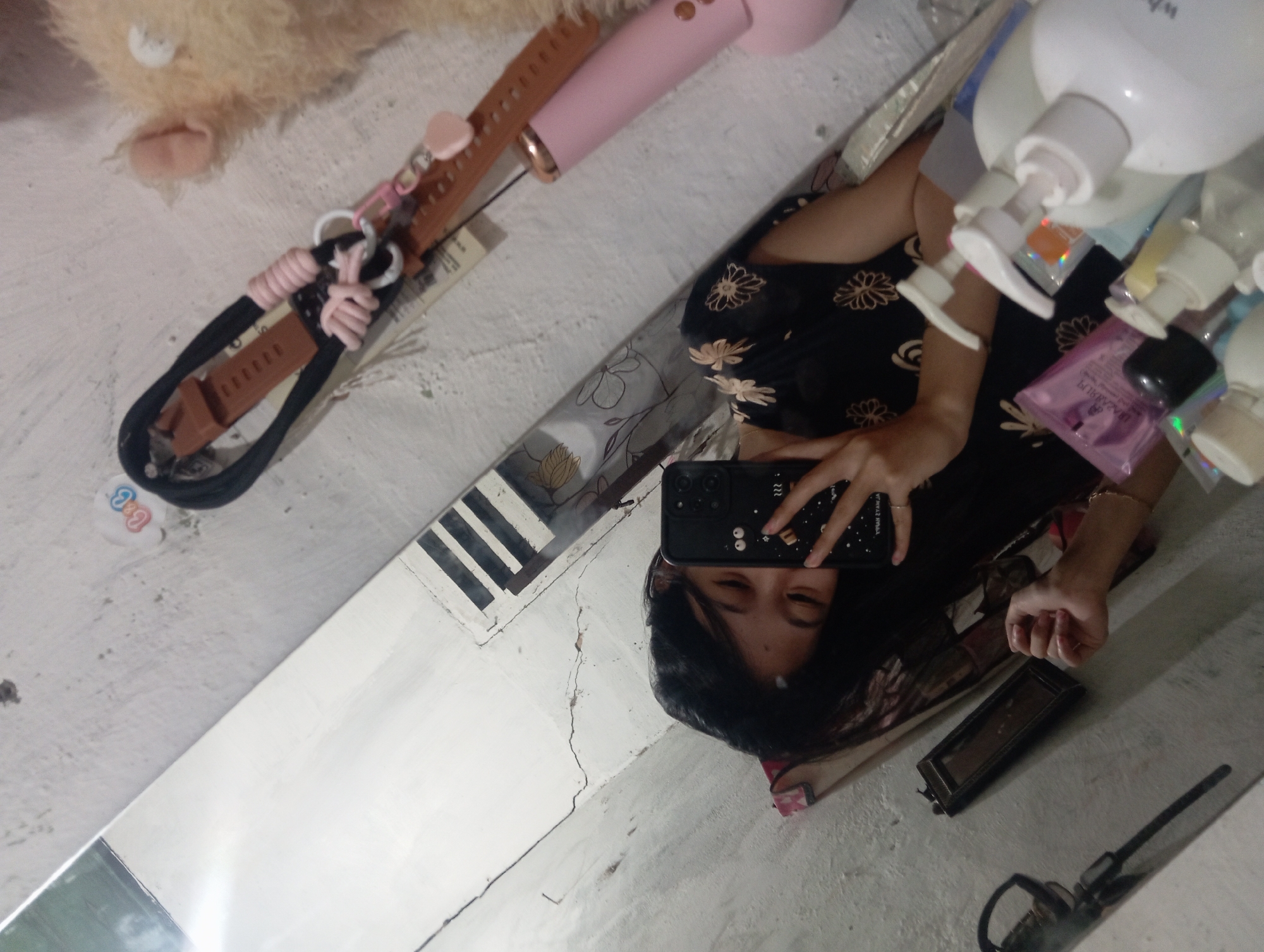 A person, appearing to be a young female with dark hair, is taking a mirror selfie in what appears to be a room in Rejosari, Indonesia. She is wearing a black floral-patterned dress. In her right hand, she holds a black smartphone with a case featuring various cartoon-like designs. The background is a cracked, light-colored wall. To the right of the person, a fuzzy, light-colored stuffed animal and a pink object, possibly a hair styling tool or electronic device, are visible on a shelf. A collection of personal care products, including pump bottles and packets, are arranged on a surface in the lower right portion of the image, partially obscured by the mirror's reflection. A thin mirror frame is also visible to the left. The image has a slightly grainy quality and is taken from a slightly elevated angle.
