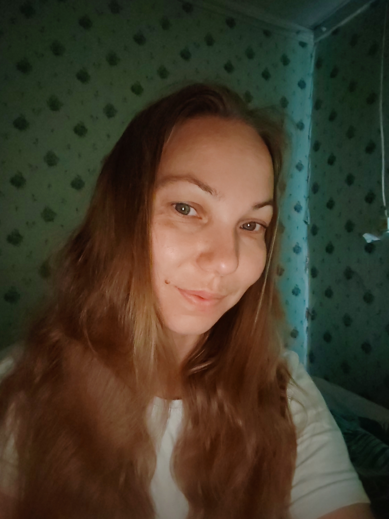 A woman with long, light brown hair is taking a selfie. She is looking directly at the camera with a slight smile. The background appears to be a room with patterned wallpaper. The image is taken indoors, likely in a residential setting. There are no visible landmarks or specific details that would allow for pinpointing a location within the city of Чухлэм, Russia.