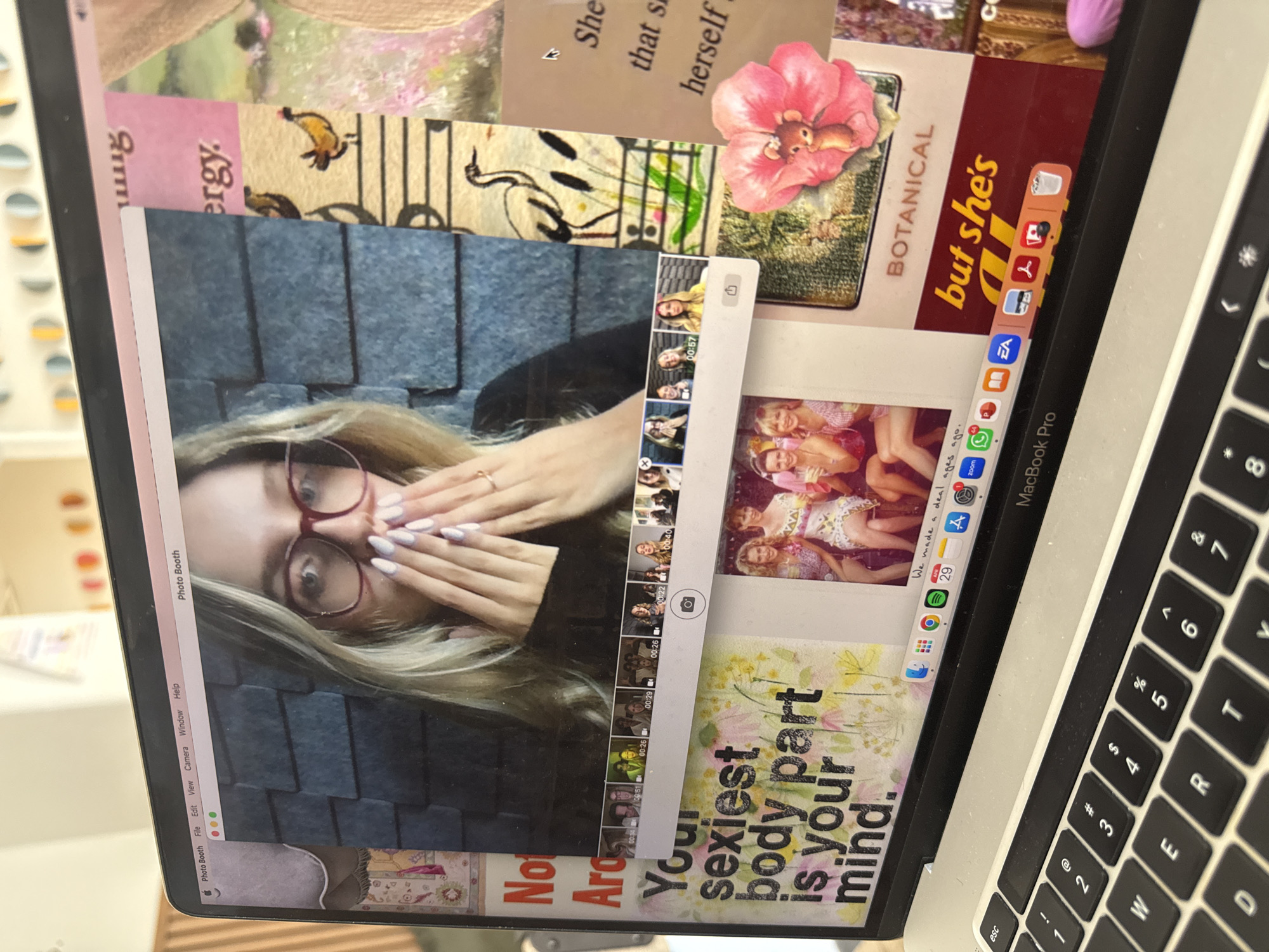 An overhead shot captures an open MacBook Pro laptop, tilted to the right, displaying a vibrant and multi-layered screen. The main subject on the screen is a young woman with long blonde hair and red-rimmed glasses, covering her mouth with her hands, which are adorned with light-colored nail polish. This image is part of a live feed from the "Photo Booth" application, whose menu bar is visible at the top.
The laptop screen functions as a digital collage, with the Photo Booth window overlaid on various background elements. These include snippets of text and images: "She that s herself," "BOTANICAL" next to an illustration of a pink flower with a tiny mouse, and "Your sexiest body part is your mind." Other background graphics feature abstract patterns and illustrations, like a bird on a musical staff. Below the Photo Booth window, a small image shows a group of young women.
The macOS dock is visible at the bottom of the screen, displaying icons for numerous applications such as Finder, Safari, Messages, Mail, WhatsApp, Zoom, Spotify, Chrome, Microsoft Word, and Adobe Acrobat, alongside others. The right side of the image shows the physical keyboard of the MacBook Pro. The background is out of focus, suggesting an indoor setting with shelves or decorative items. No specific location cues are visible within the image itself to confirm the provided context of Mexico City, Mexico. The overall impression is one of personal computer use, potentially for creative or social purposes.