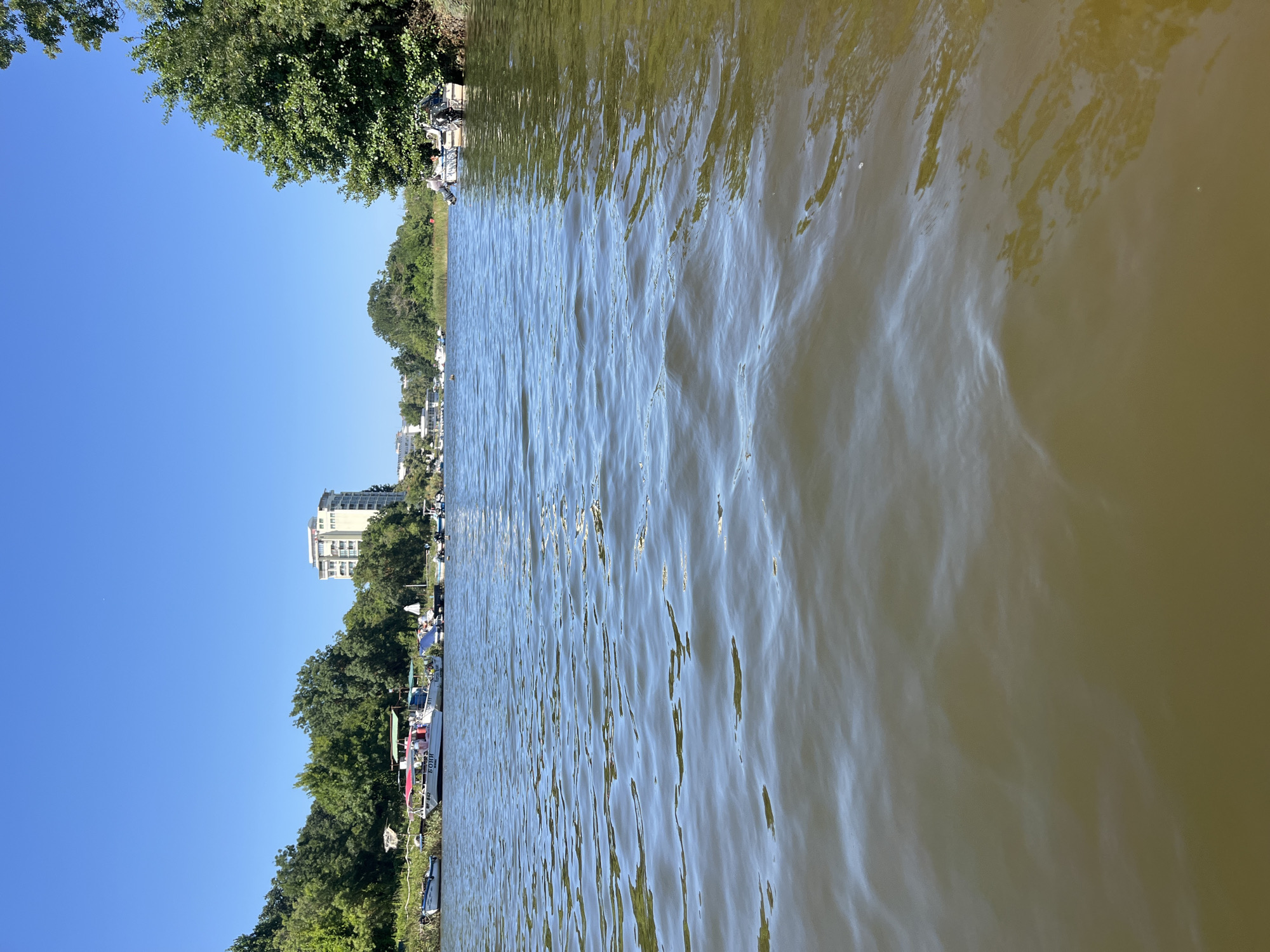 The image depicts a wide, calm river with small waves reflecting the clear blue sky. The foreground is dominated by the river's surface, showing light brown and greenish hues. Along the left bank, a line of trees and vegetation is visible, with several small boats docked. A distinctive white, multi-story building with a modern architectural style stands prominently behind the trees on the left bank. Further down the river on the right bank, more lush green trees and vegetation create a natural barrier. Two small boats, one white and one grey, with outboard motors are moored near the right bank, with at least one person visible on one of them. The overall scene suggests a sunny day in a peaceful waterside location, likely in or near Primorsko, Bulgaria, given the architectural style of the buildings and the surrounding natural environment.