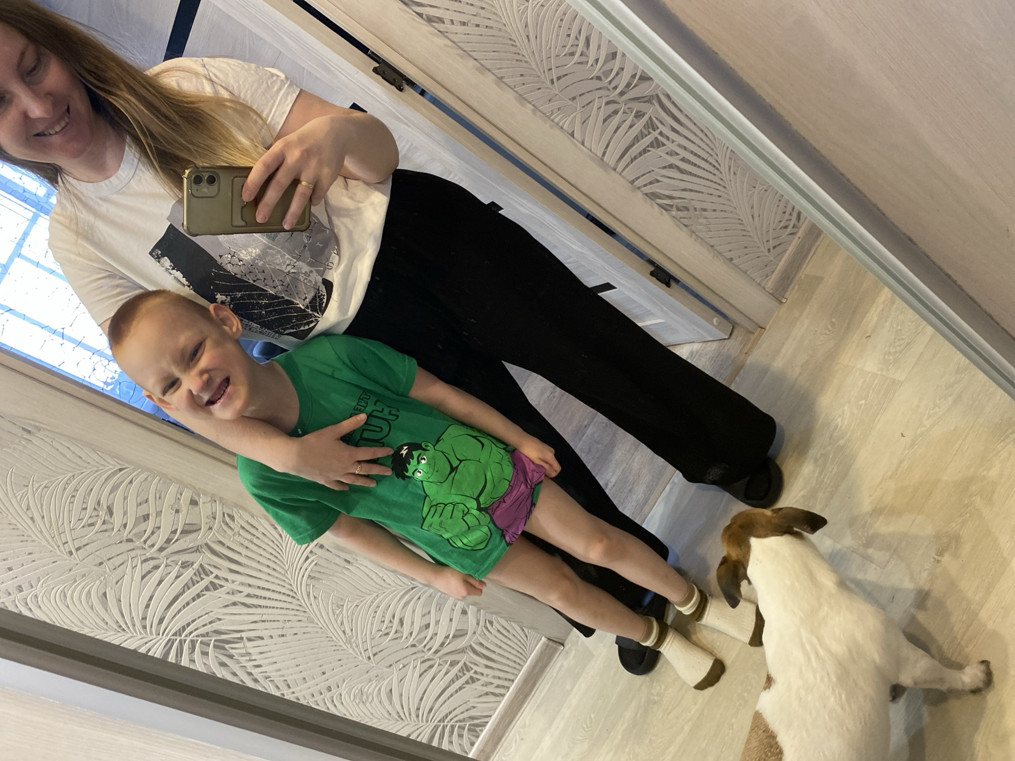 This image captures an indoor scene in Noviy, Russia, featuring a woman, a young boy, and a dog, reflected in a large mirror. The woman, with long light hair, smiles gently, looking downwards. She wears a white t-shirt with a monochrome graphic print of abstract leaves and black pants. She holds a light gold smartphone in her right hand, taking the photo, and wears a gold ring on her left ring finger. Her left arm is around the young boy. The boy, with short light hair, grins widely with scrunched eyes and an open mouth, showing a playful and happy expression. He is dressed in a vibrant green t-shirt featuring a large illustration of The Hulk, with partially visible text "THE INCREDIBLE HULK" near the shoulder, and purple shorts. He wears white socks with brown stripes near the cuff. Below the boy, a small white and brown dog looks up towards his feet. The setting appears to be a modern residential interior, possibly a hallway or room with a mirrored closet. The floor is light wood, and a wall is adorned with textured wallpaper featuring white fern or palm leaf patterns. A window on the left allows natural daylight into the space, indicating it is daytime. The overall mood is cheerful and familial.
