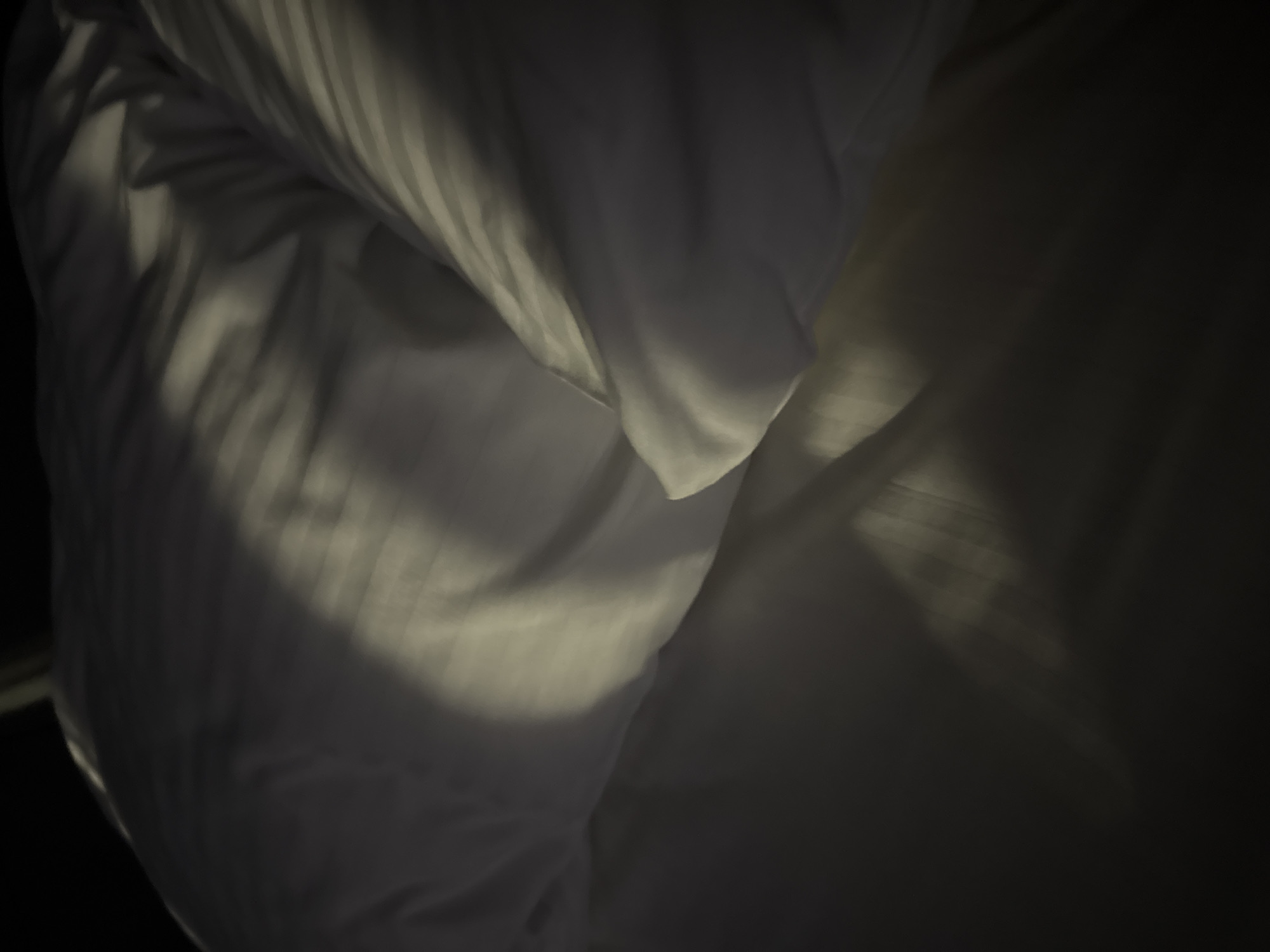 The image depicts a close-up view of a white duvet or comforter with subtle striped patterns. A diagonal shaft of light illuminates the textured surface, creating areas of highlight and shadow. The lighting suggests the presence of natural light, possibly from a window, casting distinct linear shadows across the fabric. The overall impression is one of a peaceful, undisturbed bed. The location information provided, "Doma 1096 Km, Russia," appears to be extraneous and does not correspond to any discernible elements within the image itself.