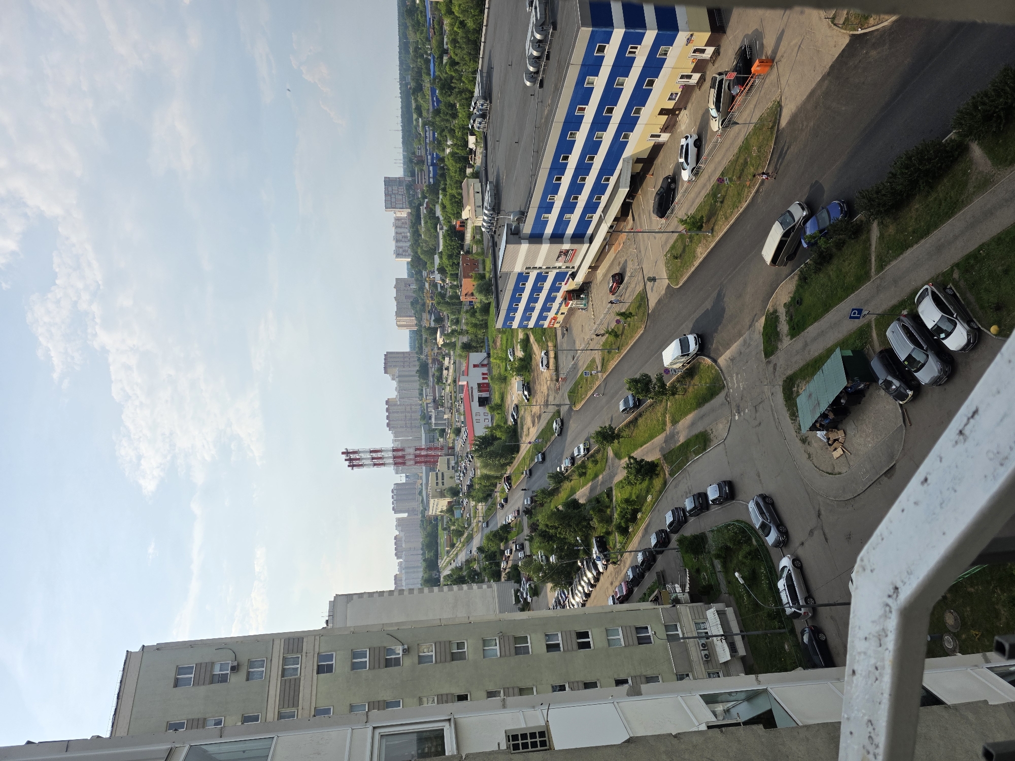 This is a high-angle, medium shot of an urban scene on a partly cloudy day. The image captures a view from a balcony overlooking a street lined with residential buildings, a large commercial building, and distant city structures.
In the foreground on the left is the side of a light-colored residential building, with several windows visible. A white metal railing runs along the bottom of the frame.
Dominating the middle ground is a road curving through the scene, with several cars parked along its sides and others driving. To the left of the road, a light green multi-story residential building is prominent, with balconies and windows. Adjacent to this building is a parking lot filled with cars.
Across the street, a large, multi-story commercial building with a distinctive blue and white striped facade stands out. Several cars are parked in front of this building, and a few individuals are walking on the sidewalk.
Further in the background, the landscape transitions to more residential buildings and industrial structures. Tall red and white striped smokestacks are visible in the distance, as well as numerous apartment blocks and green trees. The sky is a mix of blue and white clouds.
The scene depicts a typical urban environment with elements of residential living, commerce, and infrastructure. The overall impression is one of a moderately busy city on a pleasant day.