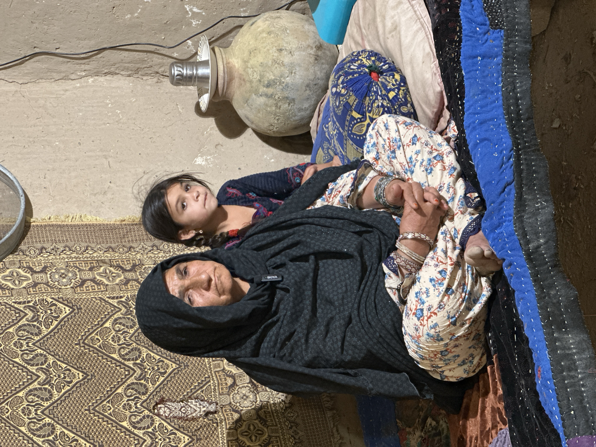 A woman and a child are lying on patterned bedding. The woman is wearing a black headscarf and floral print pants, and several bracelets. The child is beside her. Beside them are a large urn and several pillows and blankets. The location is likely Quetta City Tehsil, Pakistan.