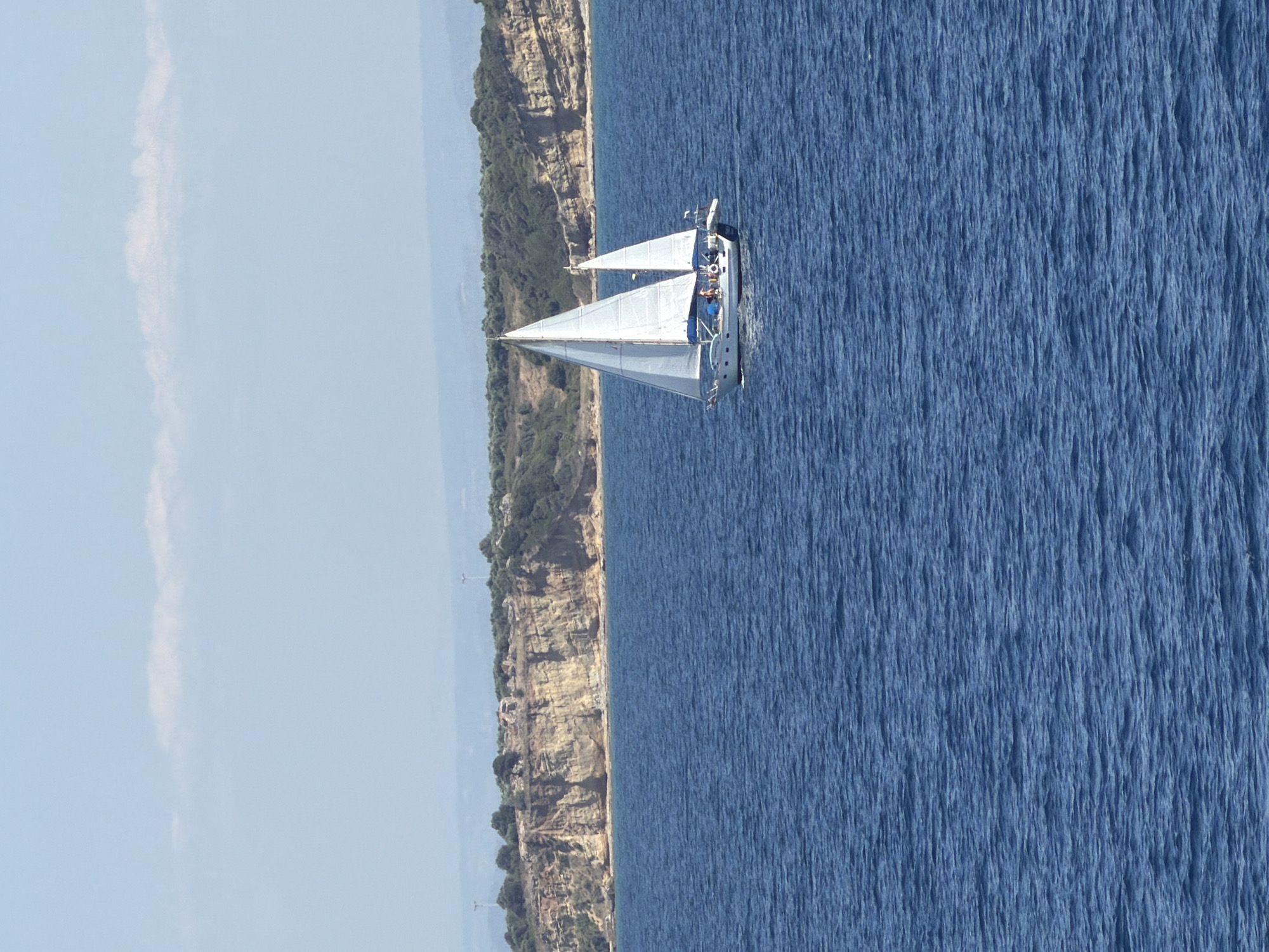 A sailboat with white sails glides across the blue water, with a rocky coastline covered in green trees in the background. The sailboat is white with a dark blue stripe along the hull, and it has two masts. Three people are visible on the deck of the sailboat. The sky is a pale blue with a thin white cloud. The scene appears to be a sunny day. The location is the Municipal Unit of Kastro - Kyllini, Greece.