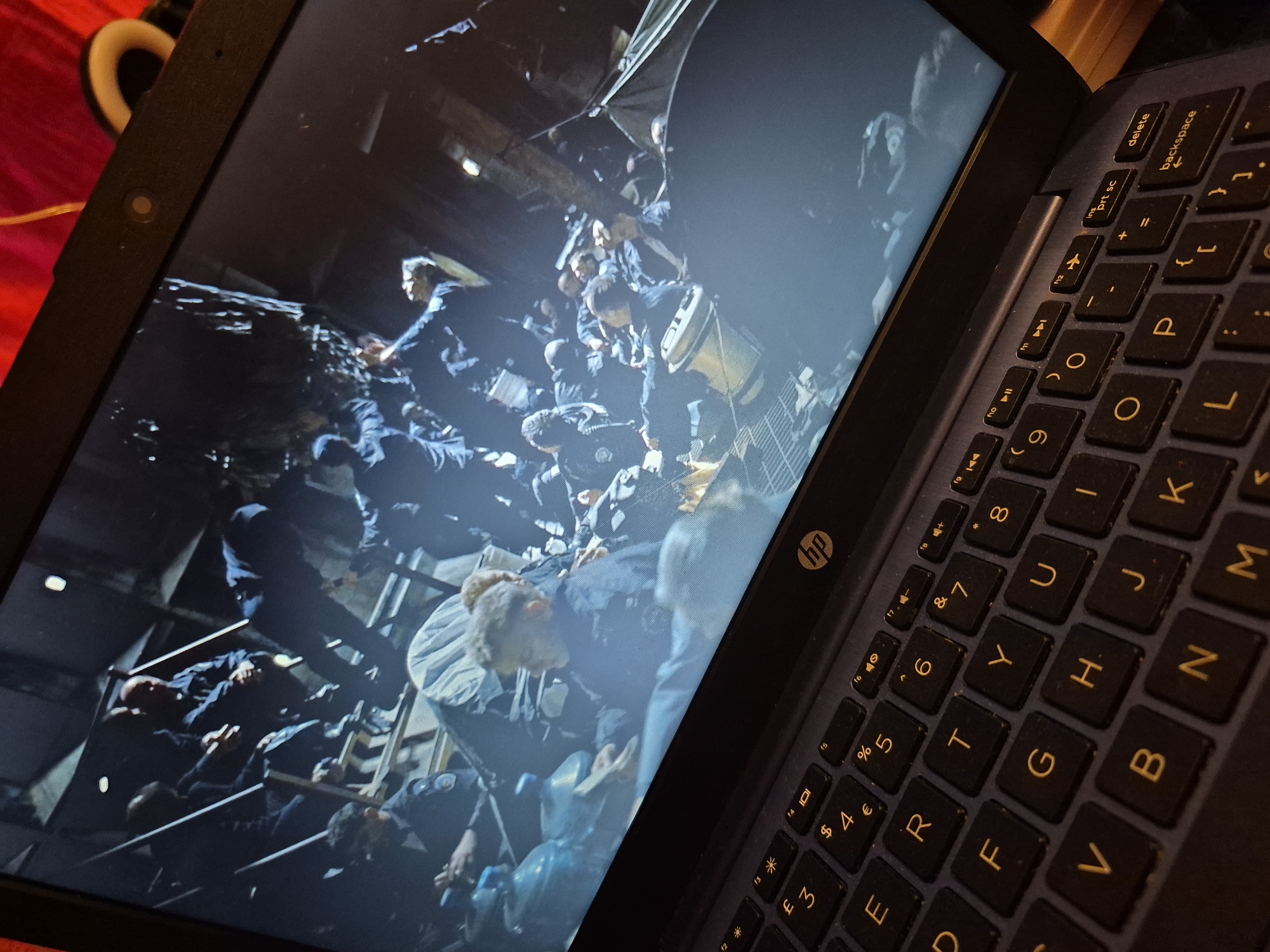 The image shows a laptop screen displaying a scene with a large group of people, some of whom appear to be wearing uniforms. The individuals are positioned on what looks like a set of metal stairs or a platform. Several people are seated, while others are standing or in motion. There are large blue containers in the foreground, possibly water bottles, and some fabric or material is visible. The overall lighting is dim, suggesting an indoor or nighttime setting. The lower portion of the image shows a close-up of a laptop keyboard, with keys like 'E', 'R', 'T', 'Y', 'U', 'I', 'O', 'P', 'F', 'G', 'H', 'J', 'K', 'L', 'V', 'B', 'N', 'M' clearly visible, along with function keys and symbols. The HP logo is present on the keyboard. The background of the image behind the laptop is blurred and shows red and white elements, potentially fabric or furniture. There is no indication of the location being in the city of Sandwell, United Kingdom.