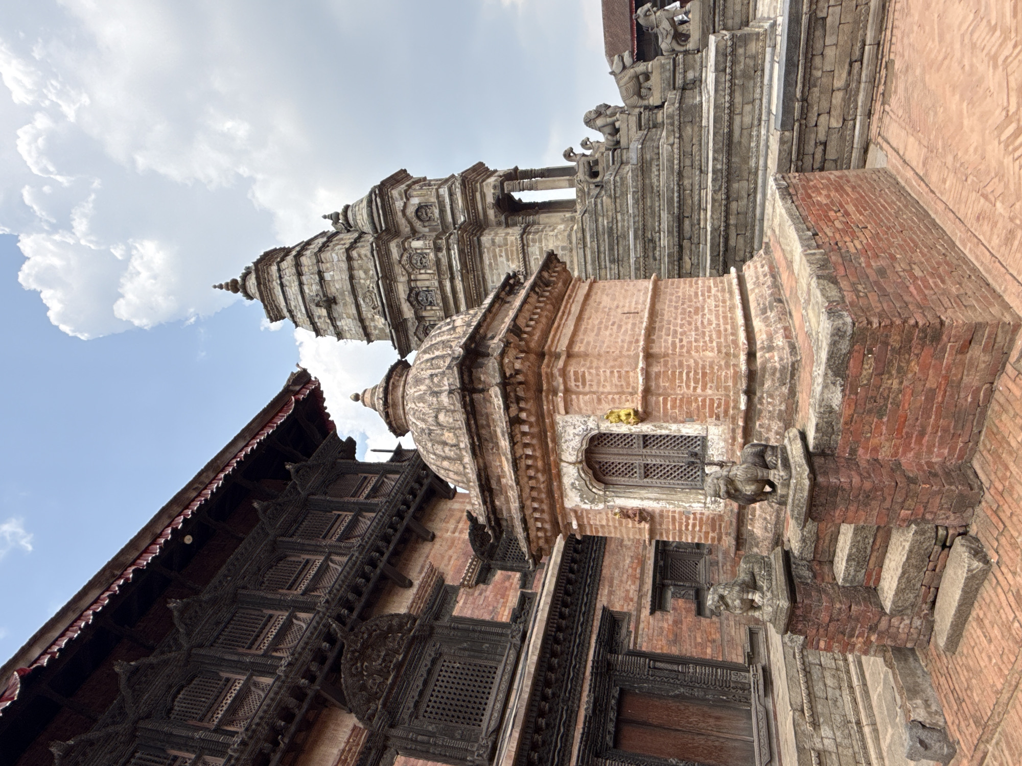 The image depicts a close-up, low-angle view of architectural structures in Bhaktapur, Nepal. The scene is dominated by several multi-story buildings and temple-like structures constructed with a combination of brick, stone, and intricately carved dark wood. The brickwork is primarily red and appears aged, while the stone elements are a lighter grey. Ornate carvings are visible on the window frames, pillars, and other architectural details. In the background, a tall pagoda-style structure rises, its layered rooflines suggestive of traditional Nepali temple architecture. The sky, featuring a mix of blue and scattered clouds, provides a backdrop.