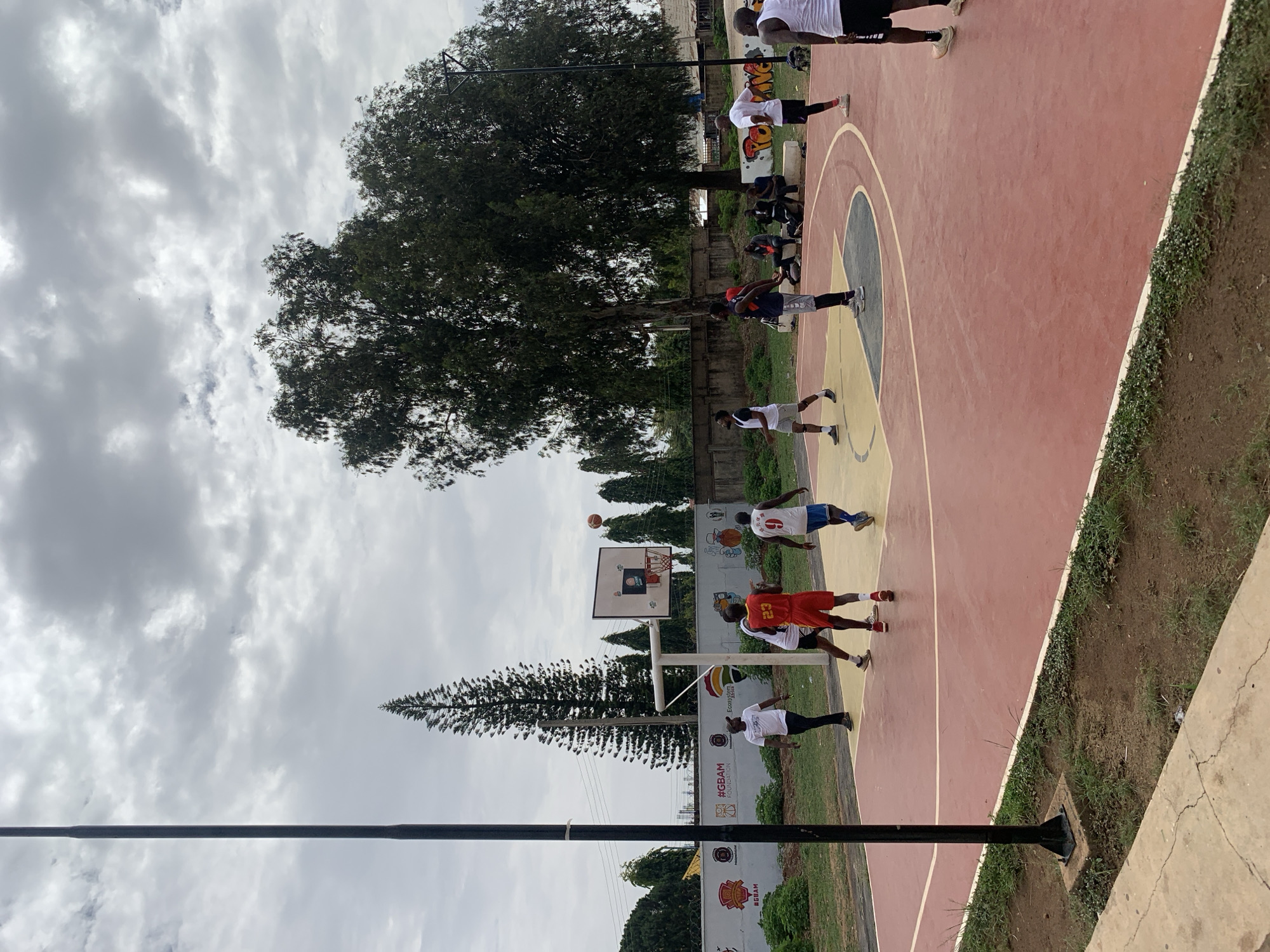 A low-angle shot of a basketball court in Gura Top, Nigeria shows several people playing basketball. The court has a red and yellow surface with white boundary lines. Players wear athletic gear, including shorts and jerseys. A basketball hoop is visible with a ball in the air above it. A large tree stands to the left of the court, and a cloudy sky covers the background. A tall, narrow conifer tree is visible near the basketball hoop. Fencing and a wall with painted advertisements flank the court. A pole is visible in the foreground, partially obscuring the view.