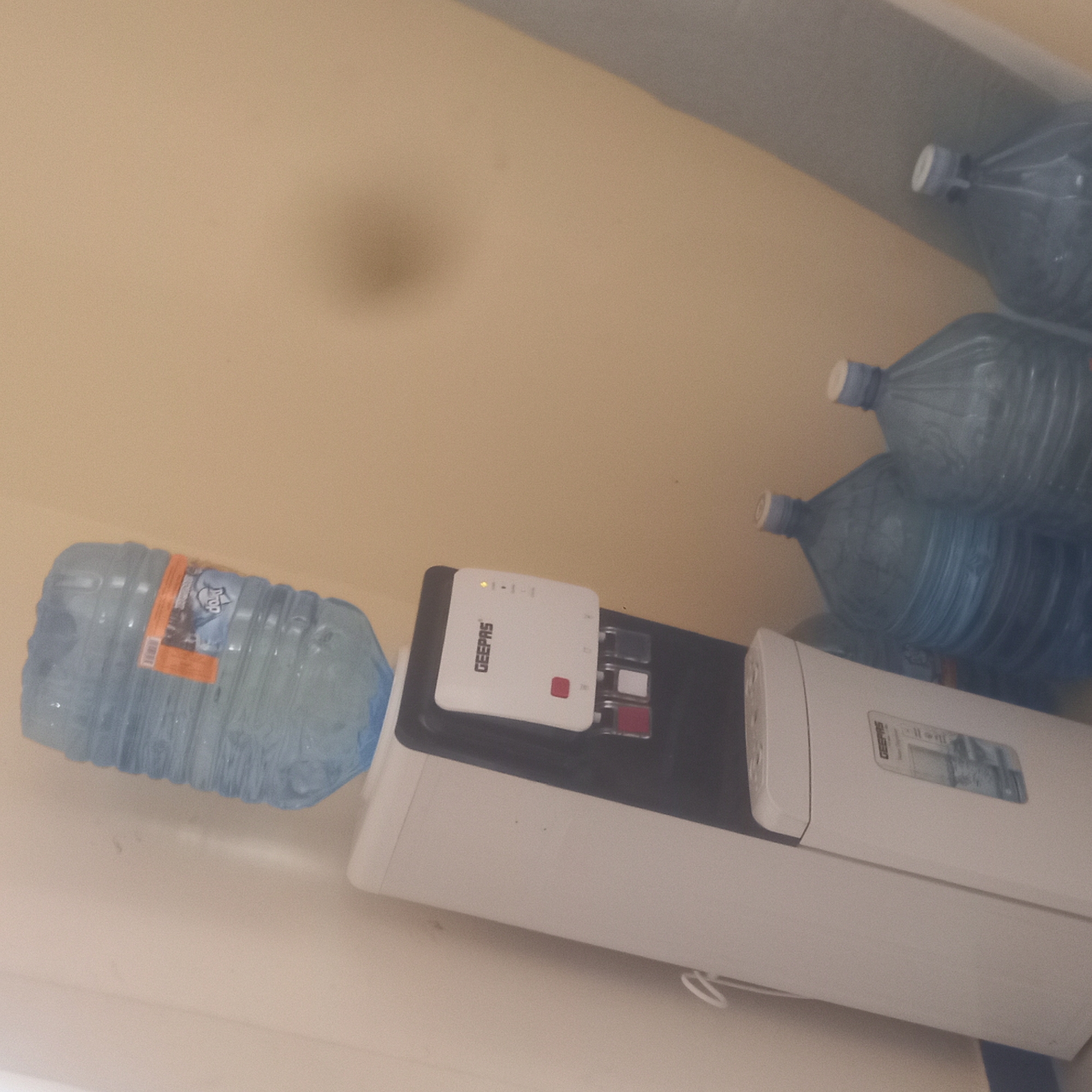 A white water cooler with a large blue water bottle on top and three blue water bottles on the floor next to it is positioned against a beige wall in what appears to be a room. The water cooler has a black panel with the brand name "GEEPAS" printed on it in white letters. Below the brand name are indicator lights and what look like dispensing taps for hot, cold, and room temperature water. A red button is visible on the left side of the panel. The water bottle on top of the cooler is partially filled, with a label that is mostly obscured but shows some text and an image. On the floor, three large, clear blue water bottles with white caps are standing upright. The environment is simple, with a plain wall and floor, suggesting a functional space rather than a decorative one. The lighting appears to be artificial and somewhat dim, casting shadows on the wall. There is a faint shadow of an object on the wall to the right of the water cooler. The image does not provide any specific clues about its location within Zanzibar, Tanzania.