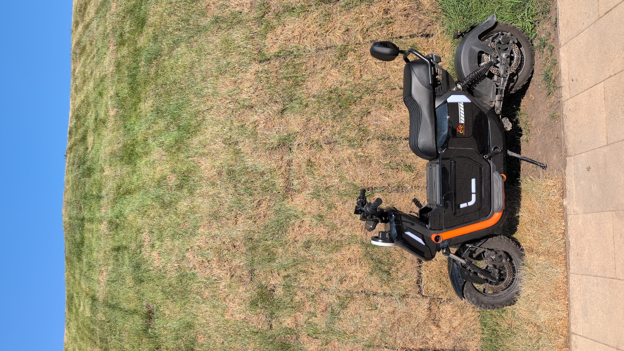 A black electric scooter with orange trim is parked on a patch of grass next to a paved walkway. The scooter has a black seat, a small backrest, and a kickstand. Behind the scooter is a steep, grassy incline that appears dry and patchy in some areas, with a clear blue sky overhead. The scene suggests an outdoor location, possibly a park or a recreational area. The image is captured in Gelendzhik, Russia, likely on a sunny day.
