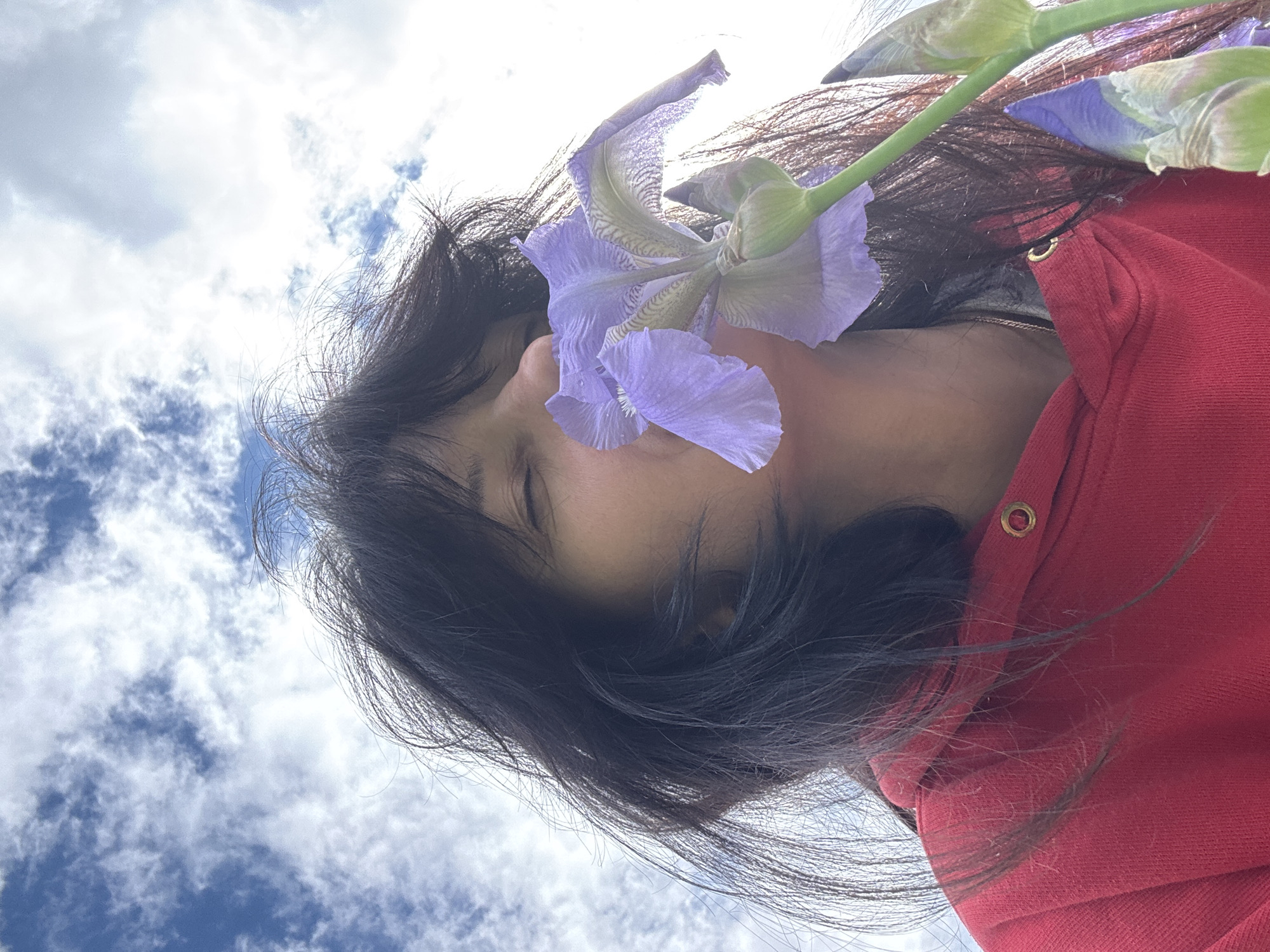 A low-angle shot shows a woman with closed eyes, her dark hair cascading towards the camera, adorned with a purple iris flower near her ear. She wears a red hooded sweatshirt with a gold button visible on the upper right. The background is a bright sky with scattered clouds. There is no information to indicate the location is in the city of Chaikovsky, Russia.