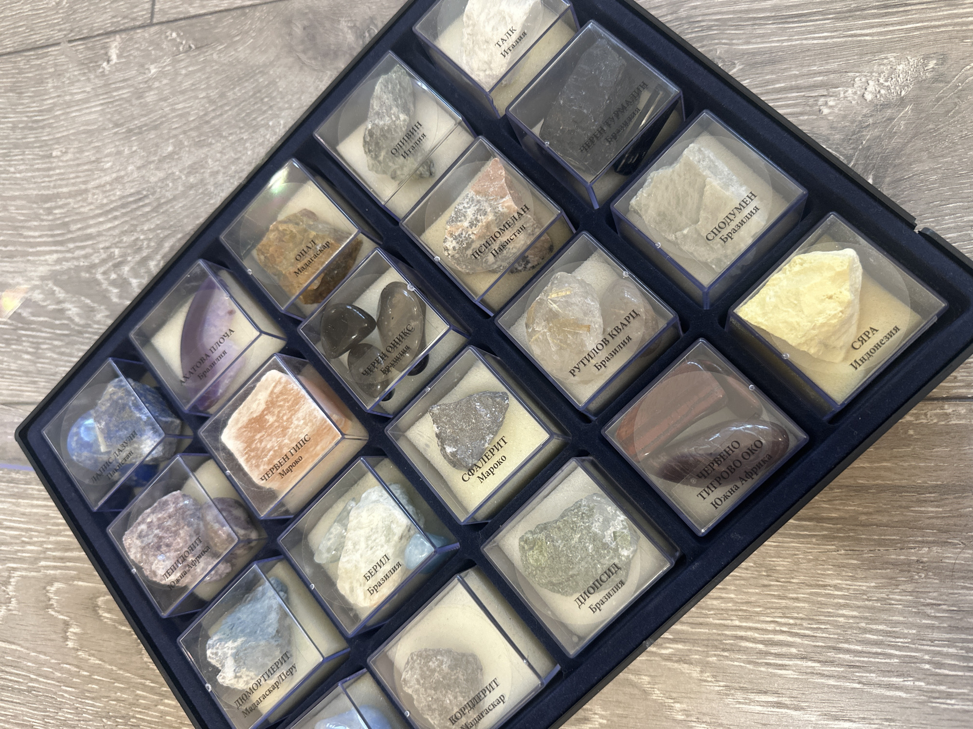 The image displays a collection of 24 mineral samples arranged in a dark blue, possibly plastic, tray. Each mineral is contained within a small, clear, square plastic box, and rests upon a white padding material. Each box also contains a small label with the name of the mineral in Russian and its origin country. The minerals vary in color, shape, and texture. The labels identify the minerals as including, among others: Talc, Olivine, Opal, Psilomelan, Rutile Quartz, Spodumene, Sulfur, Tiger Eye, Diopside, Cordierite, Beryl, and Lepidolite. The samples come from various countries, including Italy, Pakistan, Brazil, Madagascar, South Africa, Morocco, and Indonesia. The background is a light-colored wooden surface. There are no people visible in the image.
