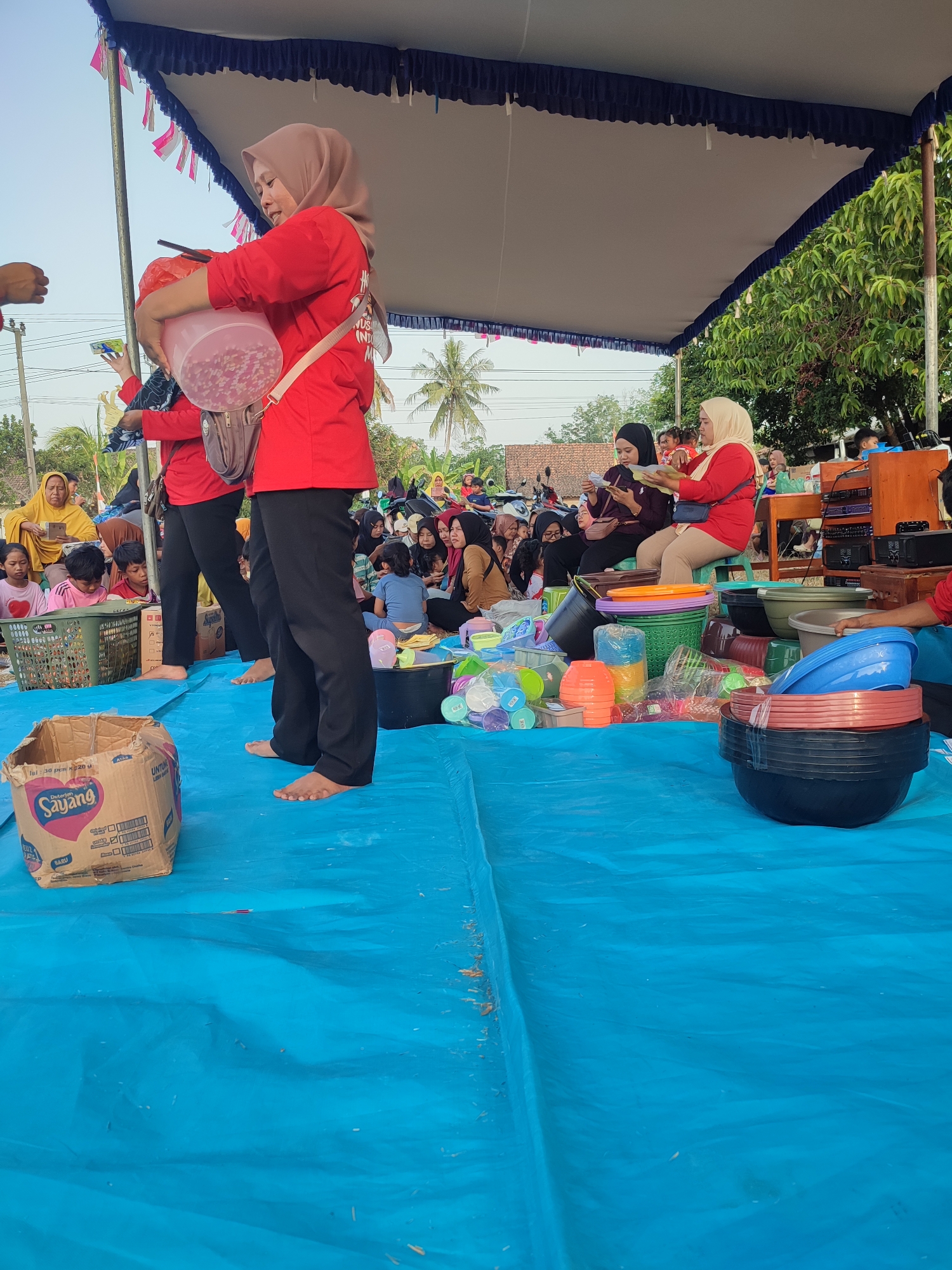 This image depicts an outdoor event in Nusa Jaya, Indonesia, likely a community gathering or market, as suggested by the presence of a tent structure, scattered audience, and various goods displayed on a blue tarp. Two women in red shirts and hijabs are at the forefront, one of whom is holding a large translucent pink container filled with small white objects, possibly prizes or candy, suggesting an activity or game is underway. Another woman in a beige hijab is seated behind them, looking at a document. A diverse crowd of people, including children and adults, are seated or standing in the background, some observing the activity. Various plastic containers and household items are laid out on the blue tarp, indicating merchandise or supplies. The environment appears to be semi-rural, with palm trees and residential structures visible in the background under a clear sky.