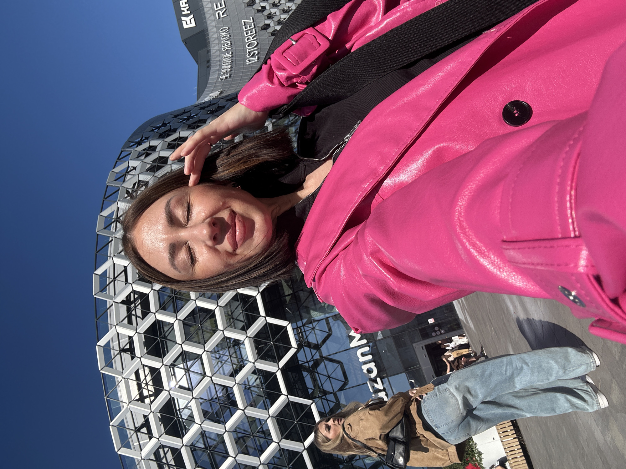 An outdoor selfie captures a young woman smiling with her eyes closed, her head tilted slightly, set against a vibrant clear blue sky and a distinctive modern building. She is wearing a striking hot pink faux leather jacket over a black top, with a black bag strap visible across her left shoulder. Her dark hair frames her face.
The background features a contemporary building with a unique facade constructed from interconnected hexagonal glass and metal panels, reflecting the blue sky. Below the main subject, another person is partially visible, dressed in a brown jacket, light blue wide-leg jeans, and white sneakers, carrying a black shoulder bag. This individual appears to be standing or walking near the building's entrance.
The scene is brightly lit by natural daylight, indicating a sunny day. Visible text on the building includes "KAPP", "ZOLOTOE YABLOKO" (Golden Apple), "12STOREEZ", and partially "RE" and "Kazan", suggesting an urban commercial area within Kazan, Russia. The ground is a paved surface, and blurry figures are visible near the building's entrance in the background. The overall mood is cheerful and relaxed.