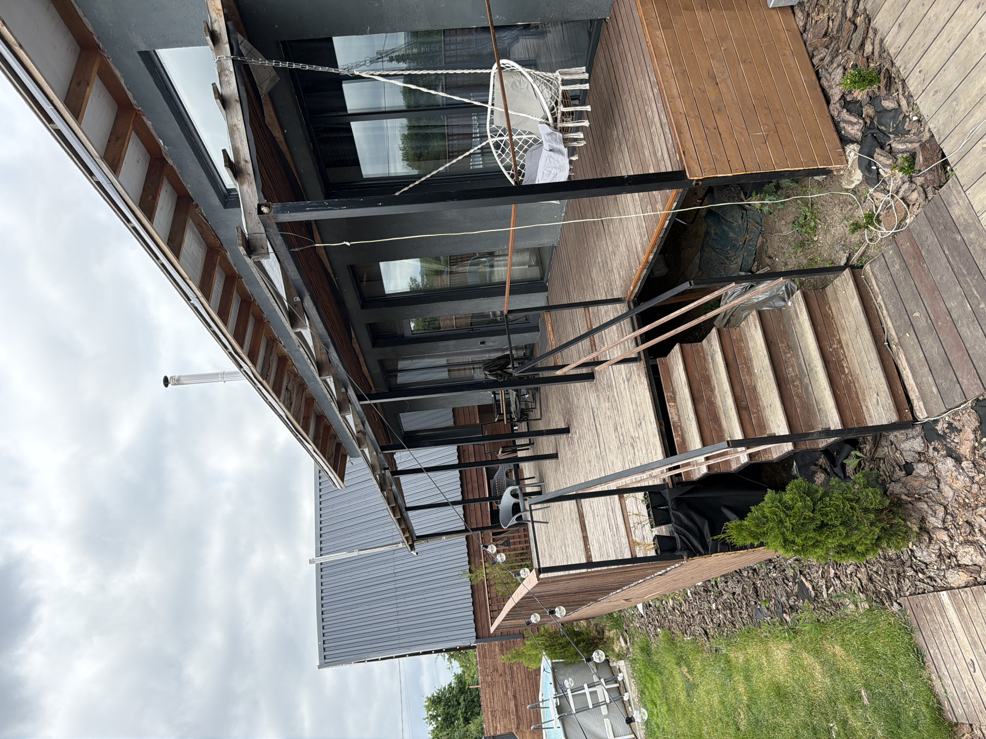 The image depicts the exterior of a modern house with a wooden deck and a covered porch in the city of Semiluki, Russia. The house has a gray facade and large windows. On the deck, there is a white hanging swing chair with cushions, several chairs and tables, and string lights. Stairs lead down from the deck to a wooden walkway. To the left of the deck, a small above-ground pool is visible, surrounded by grass and bark mulch. In the background, another building with a gray, corrugated metal roof is present. The sky is overcast with gray clouds. The overall scene suggests a residential property, possibly a vacation home or a modern dwelling.