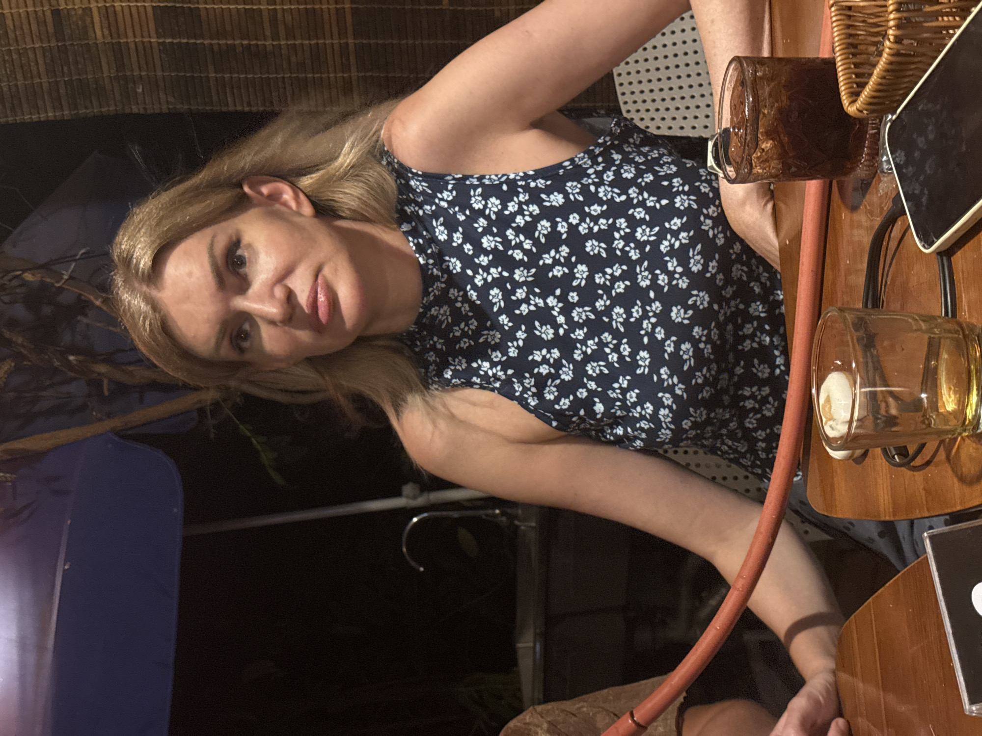 A blonde woman in a navy blue dress with a white floral pattern is seated at a wooden table at night. She is looking to her left. In front of her is a hookah hose, a glass with a brown beverage and ice, a smaller glass with a yellowish liquid, and a cell phone. To her left, a person's arm and shoulder are visible. Behind the woman, a woven bamboo blind is visible. The environment appears to be an outdoor seating area, possibly a restaurant or bar, in Nha Trang, Vietnam.