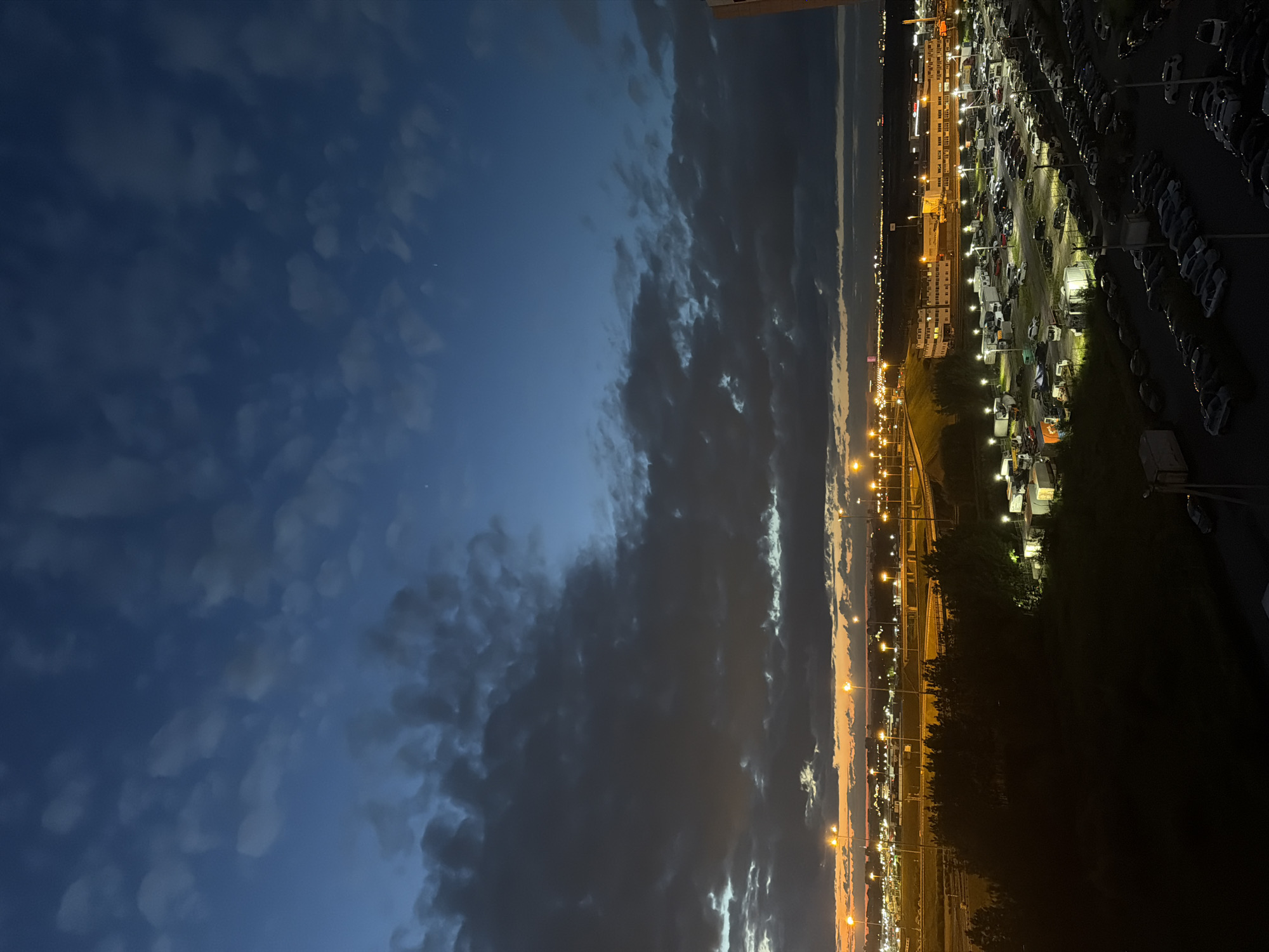 The image shows a dusk or twilight scene above a large, illuminated parking lot filled with many vehicles. The sky is a deep blue, with scattered dark clouds and some lighter patches near the horizon where the sun has recently set, casting an orange glow. Numerous streetlights illuminate the parking lot and the surrounding infrastructure, including roads and buildings in the distance. The foreground is dark, suggesting the vantage point is from a higher elevation, possibly a building. There are no people clearly visible. The overall impression is of an urban or industrial area at the end of the day.