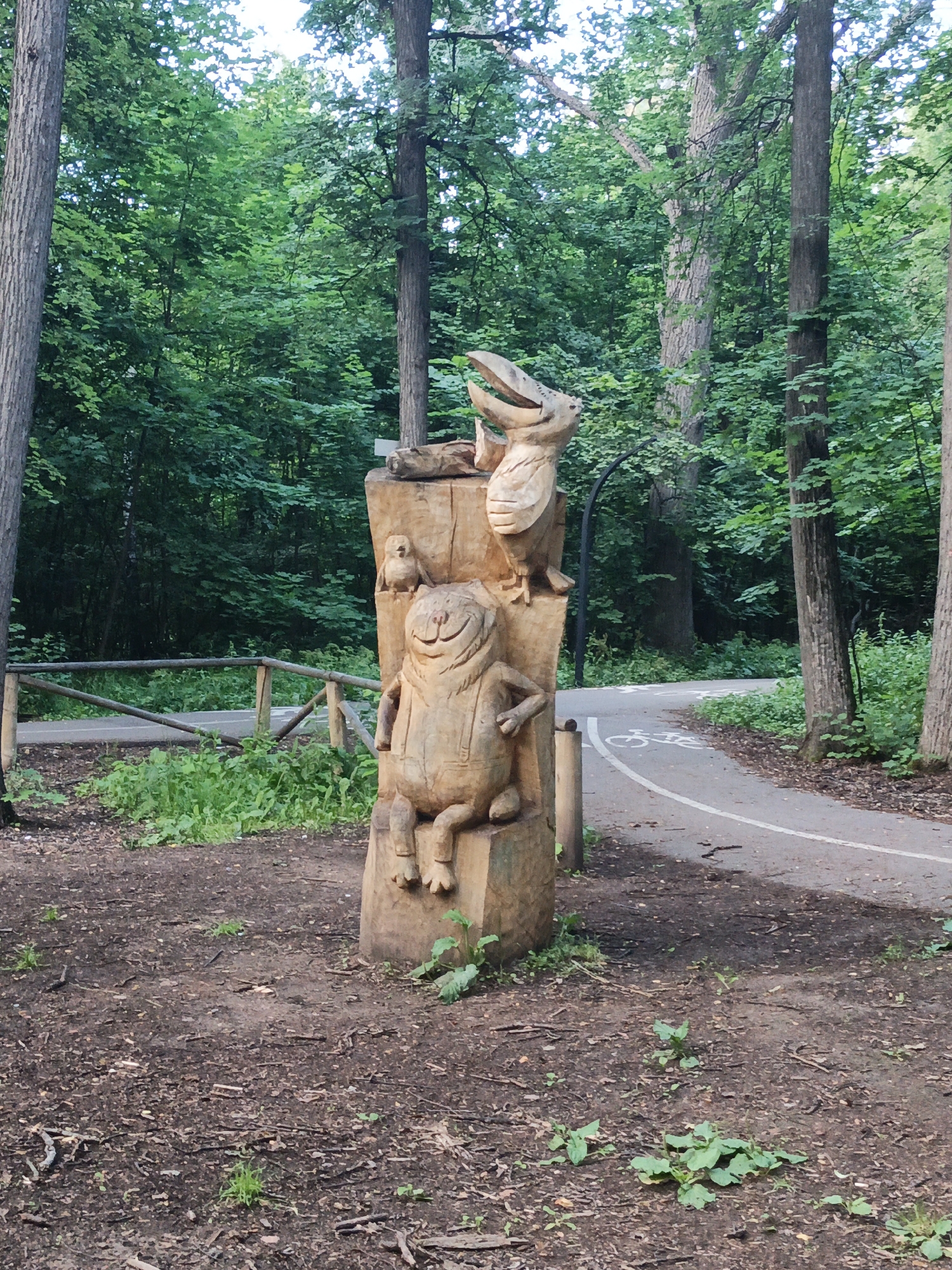 This media file shows a whimsical wooden sculpture of cartoonish animals in a park setting. The main subject is a carved wooden figure that appears to be a cat or frog-like creature, sitting with a jovial expression, wearing overalls. Above it, a bird-like creature with its mouth open is carved into the same wooden pillar. A smaller bird is perched on the pillar to the left of the larger bird. The sculpture is set outdoors amongst trees, with a paved path for cycling visible in the background, indicated by a bicycle symbol painted on the asphalt. A wooden fence runs along the edge of the path. The ground is covered in dirt, leaves, and scattered twigs. The scene is lush and green, suggesting a warm day, possibly in late spring or summer, with sunlight dappling through the trees. The overall impression is one of a peaceful, natural environment enhanced by playful art. The location is identified as Ilteryakovo, Russia. There is no visible text in the image.