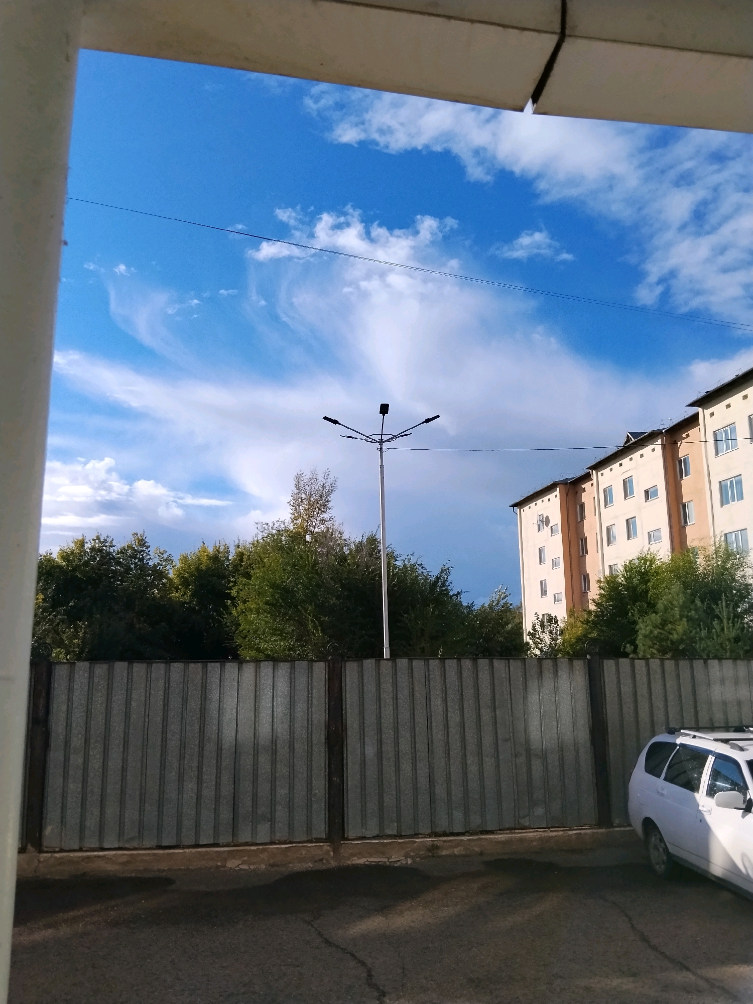 The image captures a street scene with a multi-story residential building and a metal fence in the foreground. The sky is blue with scattered white clouds. A street lamp with multiple heads stands near the center of the frame. Trees are visible behind the fence. A white station wagon is parked on the right side of the frame. The scene appears to be in an urban or suburban area, likely Kokshetau, Kazakhstan, given the presence of typical Soviet-era apartment blocks. The time of day seems to be daytime, possibly late afternoon or early evening, with the sun casting shadows on the ground. There are no people visible in the image. The environment is characterized by residential architecture, natural elements like trees, and man-made infrastructure such as the fence and street lamp.