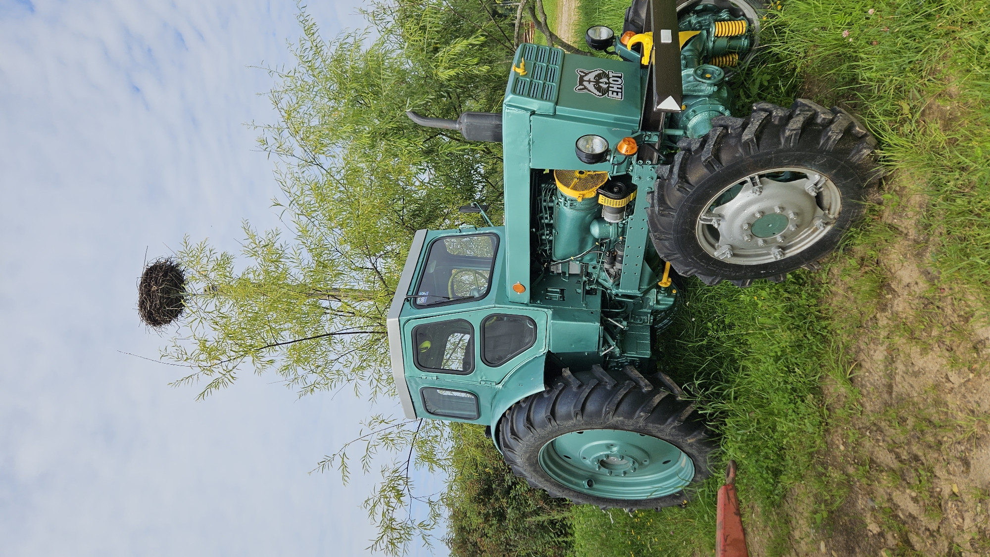 This image depicts a vintage, teal-colored tractor parked on a grassy area in Kapūņi, Latvia. The tractor has large, rugged tires and a visible engine with yellow accents. A distinctive raccoon decal with the word "EHOT" is affixed to the front of the tractor. In the background, a tall tree supports a large bird's nest at its apex. The sky is partly cloudy, with white clouds against a light blue backdrop. The overall environment suggests a rural or semi-rural setting.