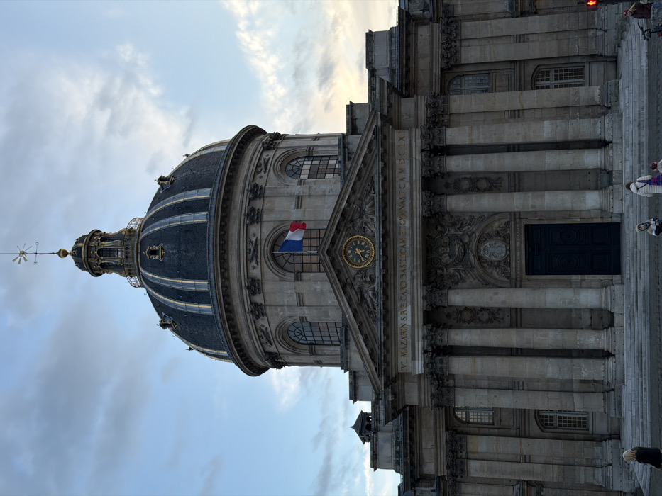 The image showcases a grand building, possibly a church or state building, under a cloudy sky. The architecture features a large, ornate dome with a spire. A French flag hangs from a window. Below, a detailed façade includes columns, a clock, and an inscription, leading to a large door. People are present on the steps, adding a sense of scale. The image's perspective is slightly tilted.